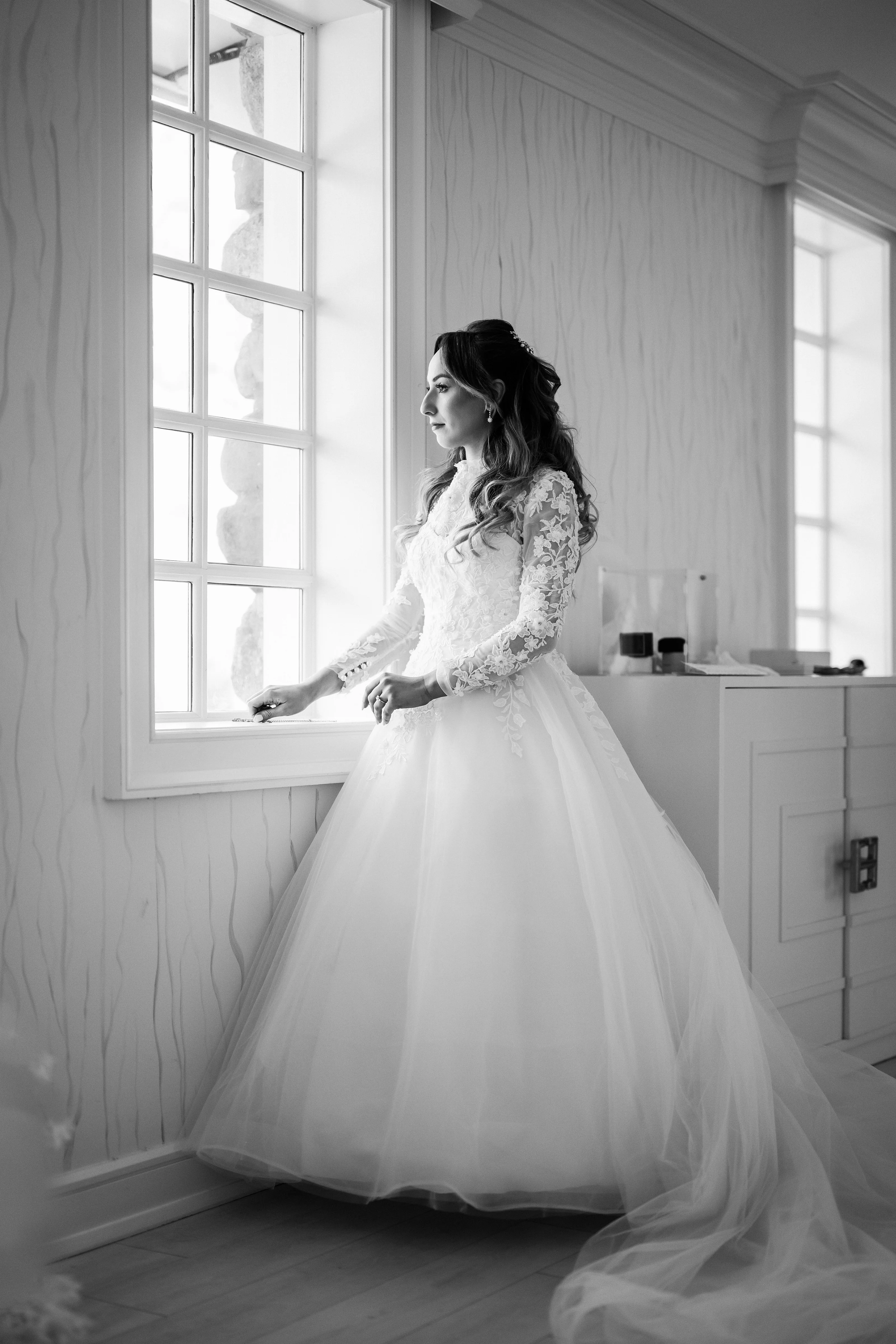 Black-and-white portrait of the bride in a lace gown standing by tall window light, turned in profile with a calm expression.