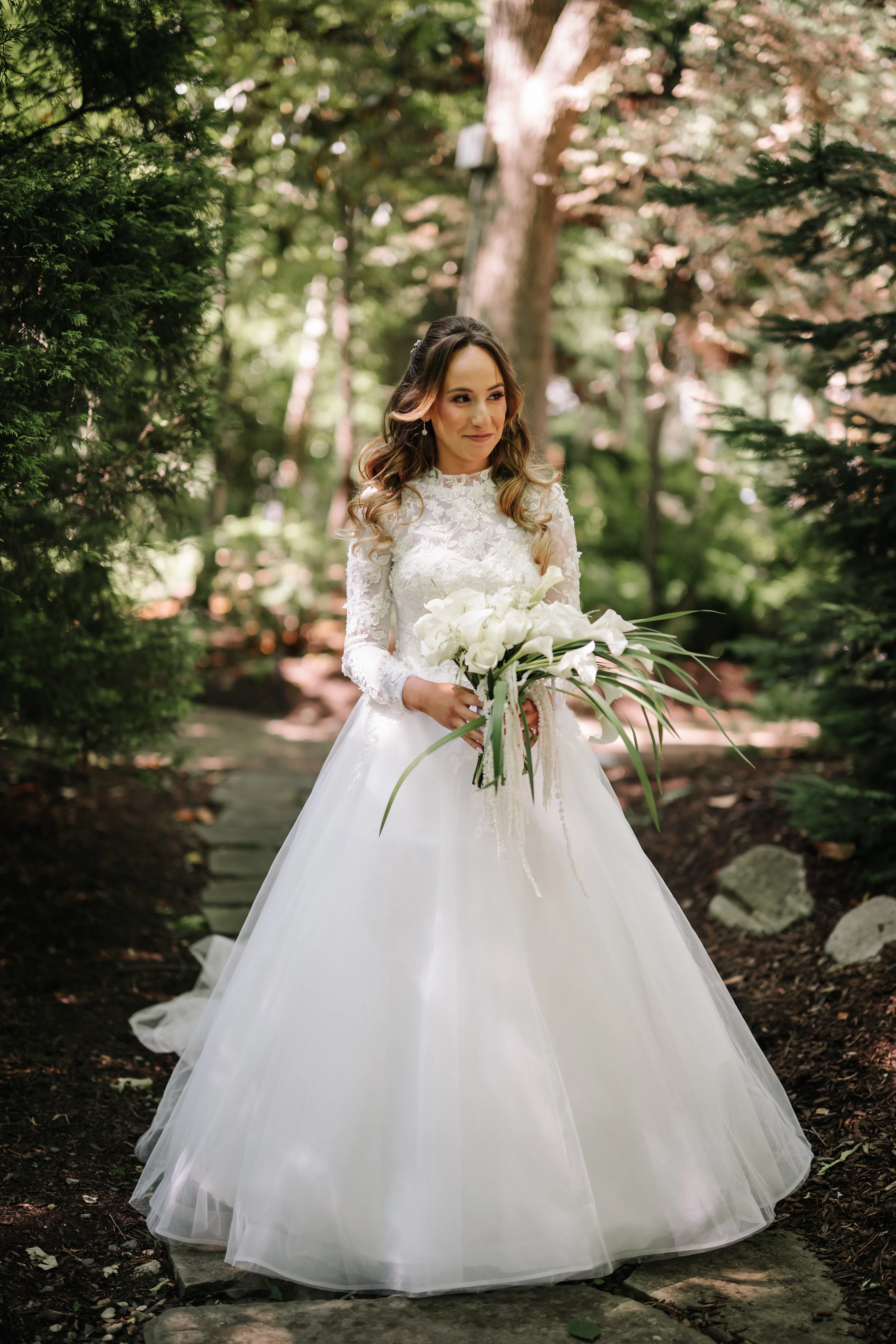 Bride holding a white bouquet outdoors in a garden with soft dappled light through trees.