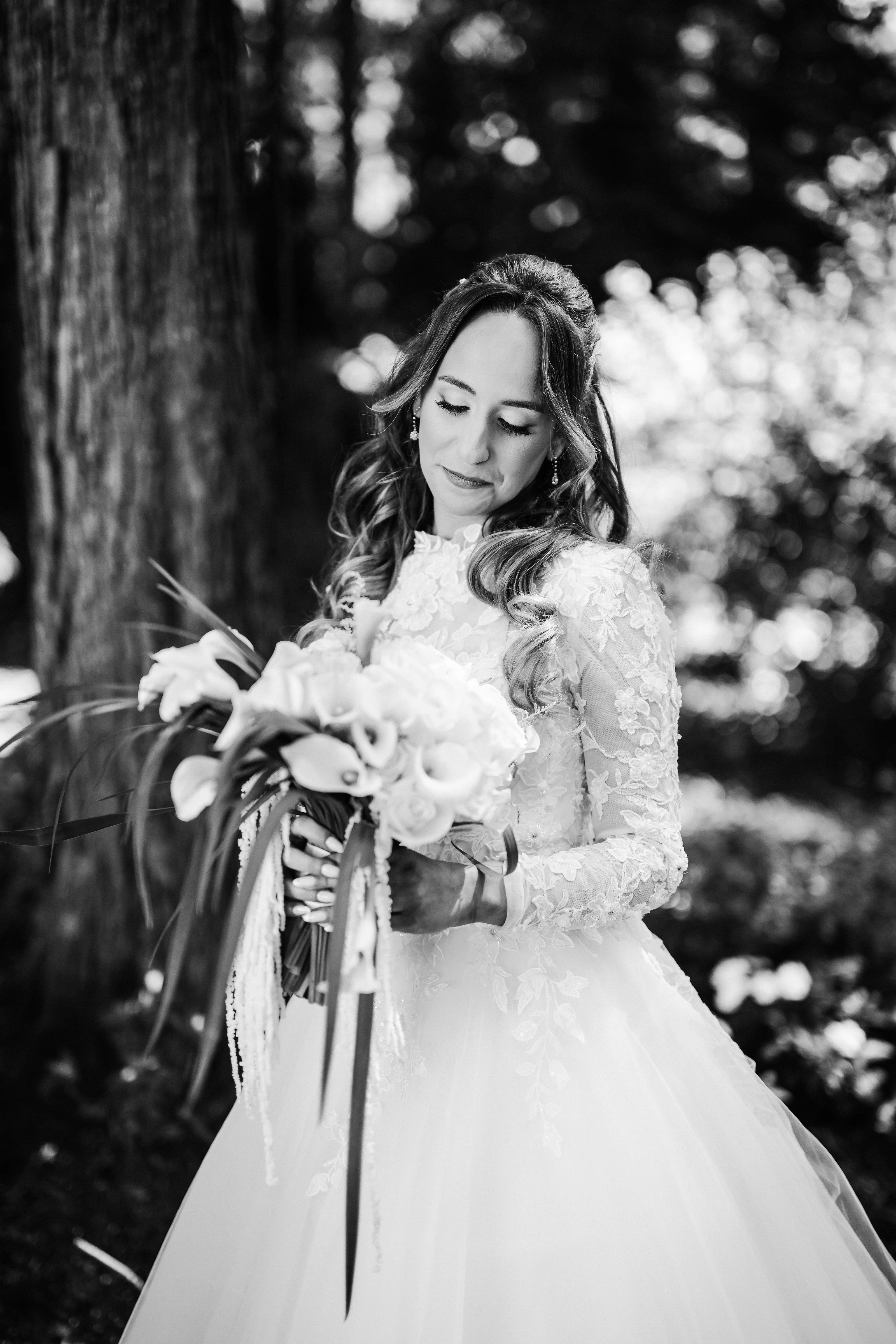 Black-and-white wedding portrait of the bride holding a bouquet with her eyes lowered, framed by a softly blurred outdoor garden background.