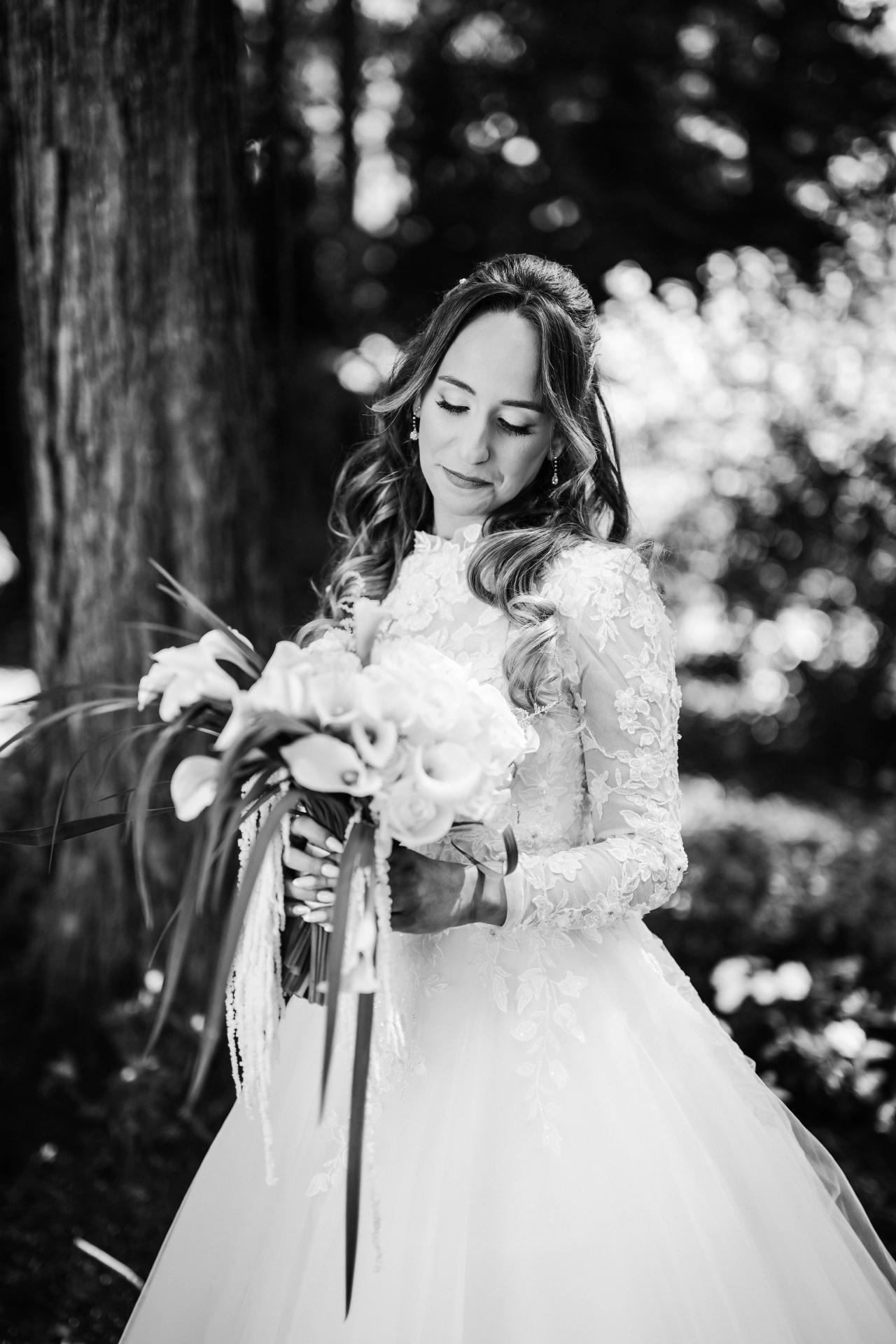 Black-and-white wedding portrait of the bride holding a bouquet with her eyes lowered, framed by a softly blurred outdoor garden background. Wedding at Crystal Plaza, Livingston, NJ.