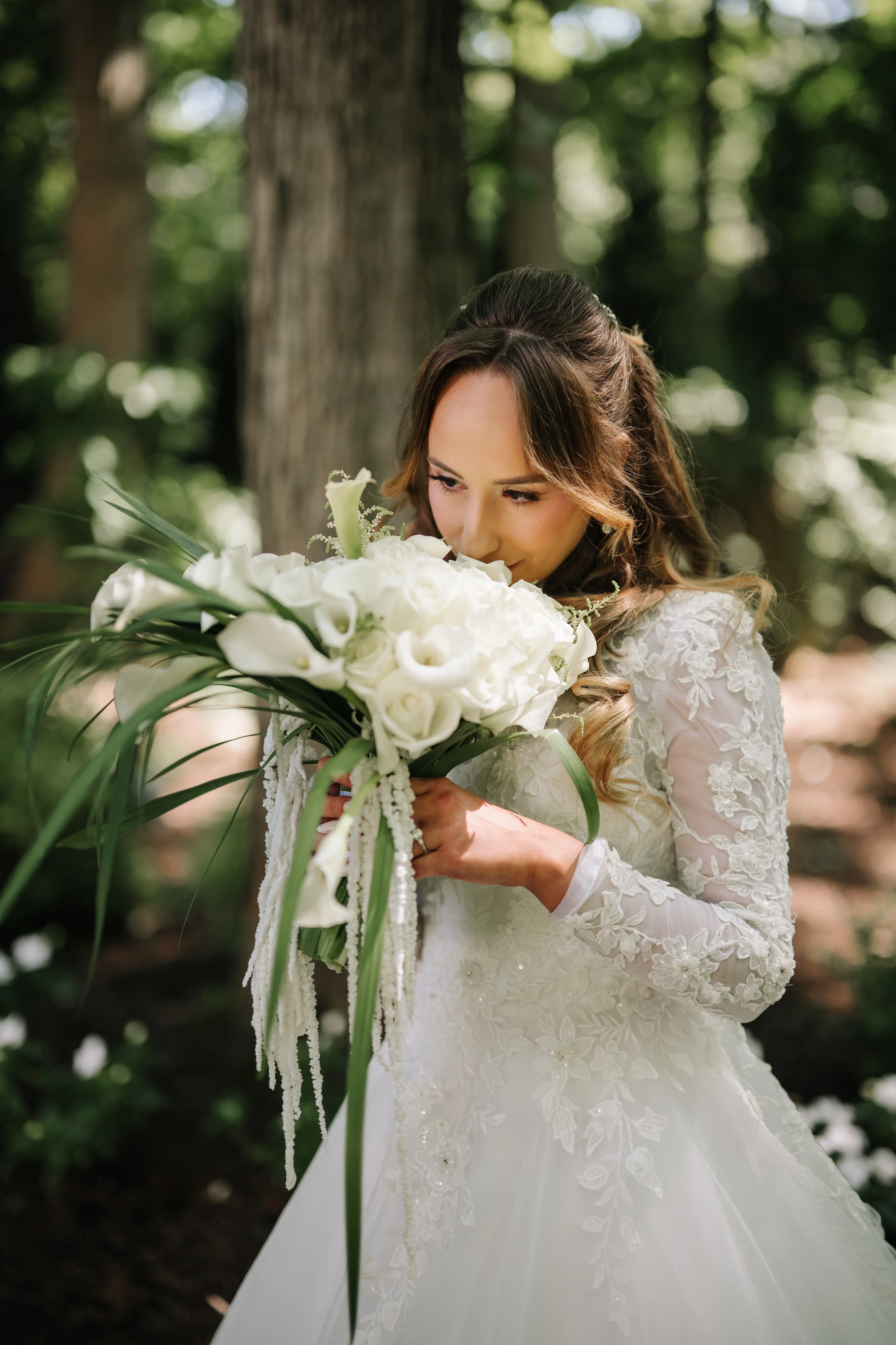Brunette bride in a lace dress holding a white bouquet close, eyes down, in warm outdoor greenery with soft bokeh.