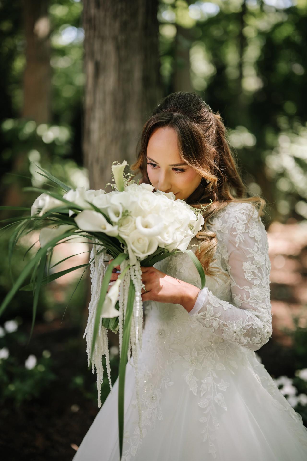 Brunette bride in a lace dress holding a white bouquet close, eyes down, in warm outdoor greenery with soft bokeh. Wedding at Crystal Plaza, Livingston, NJ.