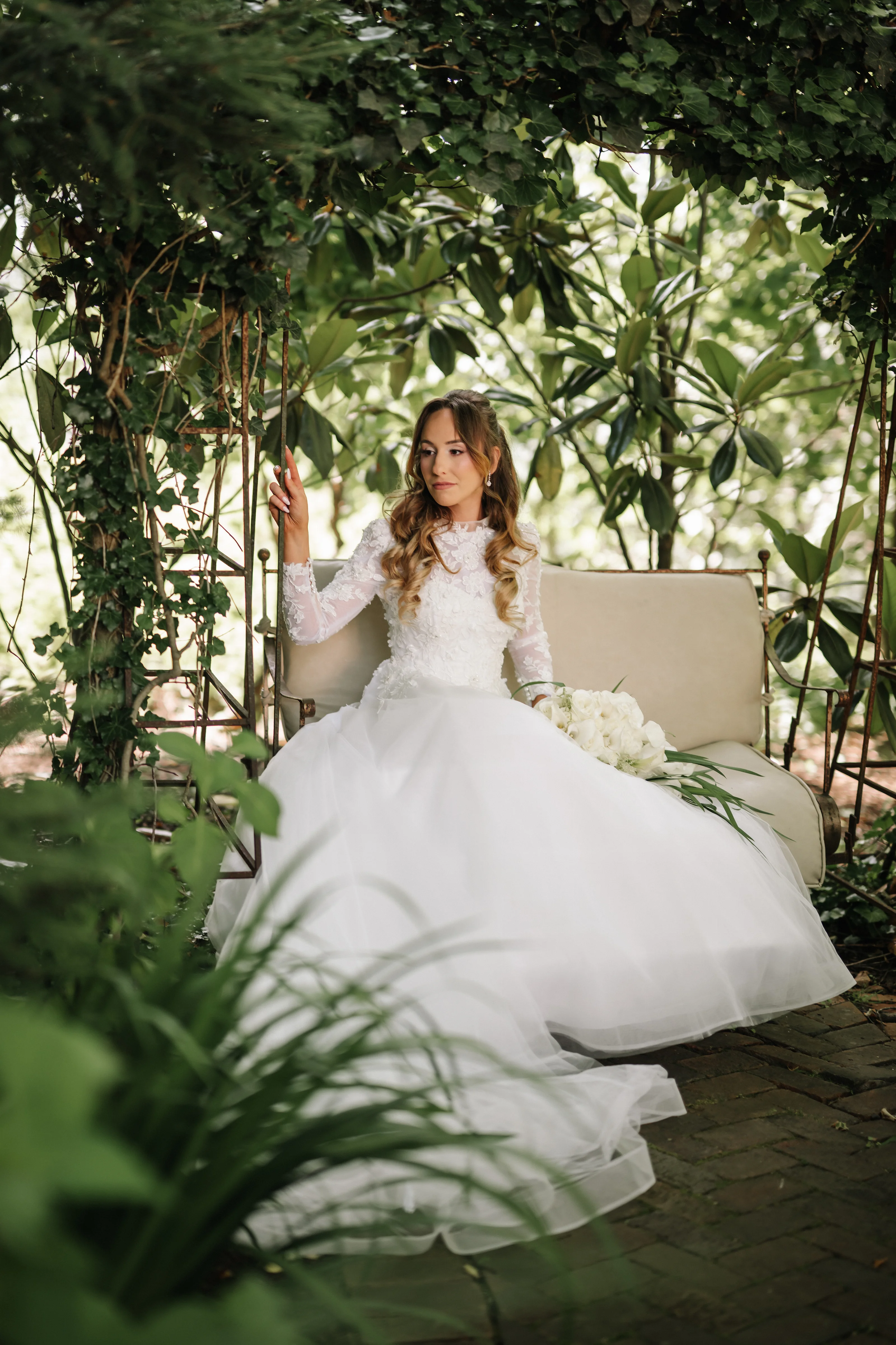 Bride in a white wedding gown seated on a garden lounge chair framed by lush greenery, softly lit through leaves.