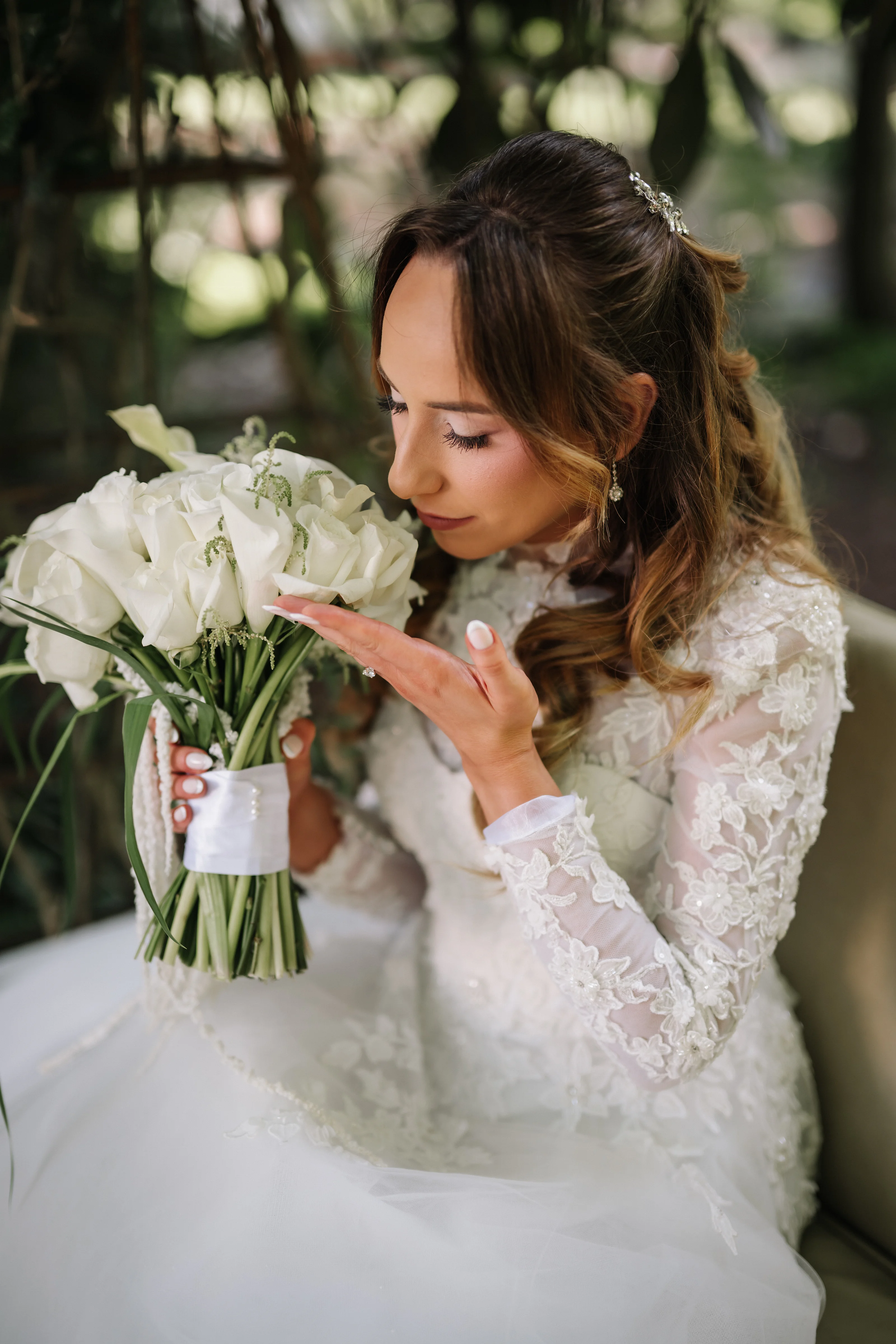 Bride in lace attire holding a white bouquet, eyes closed, with soft garden light and a blurred green background.