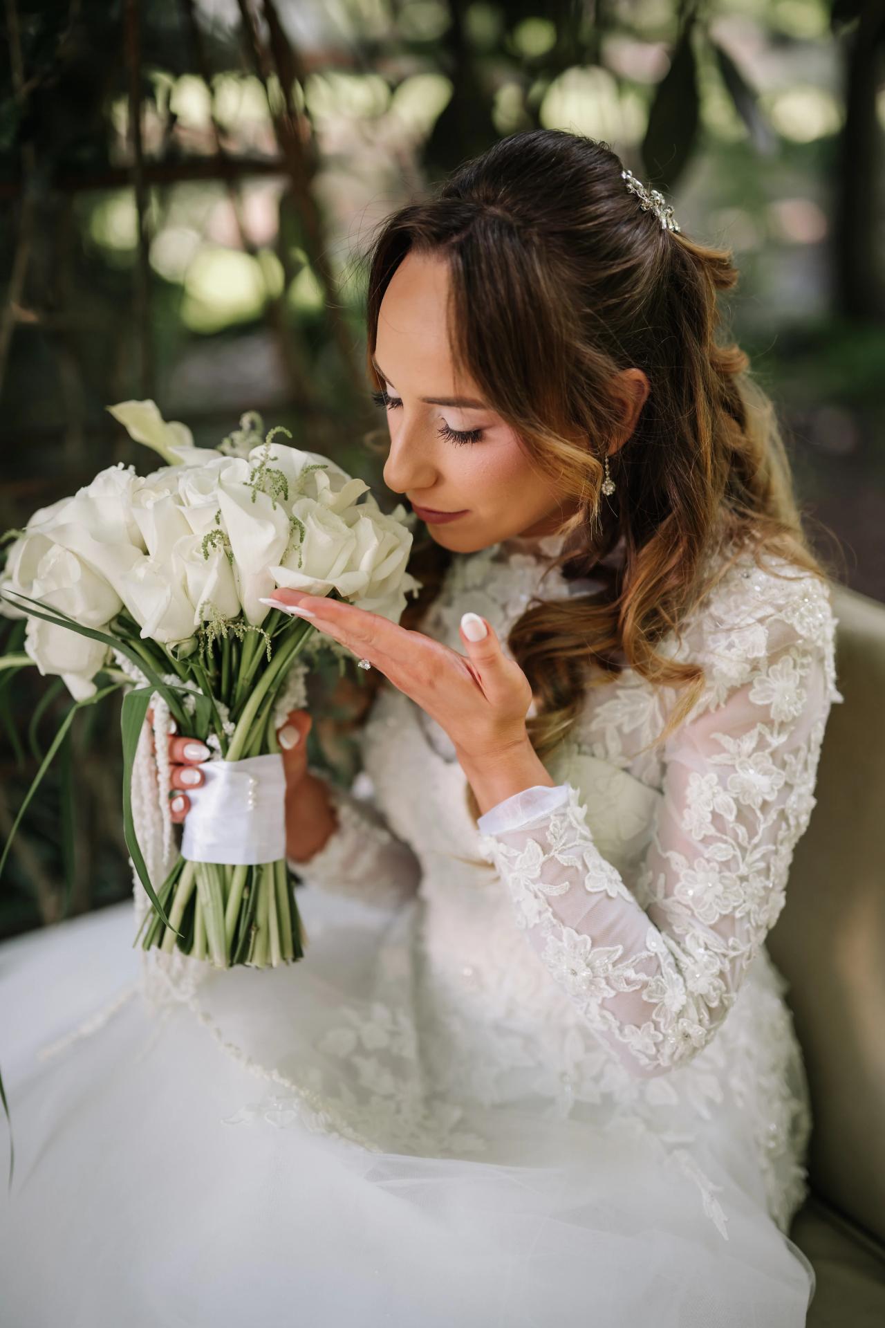 Bride in lace attire holding a white bouquet, eyes closed, with soft garden light and a blurred green background. Wedding at Crystal Plaza, Livingston, NJ.