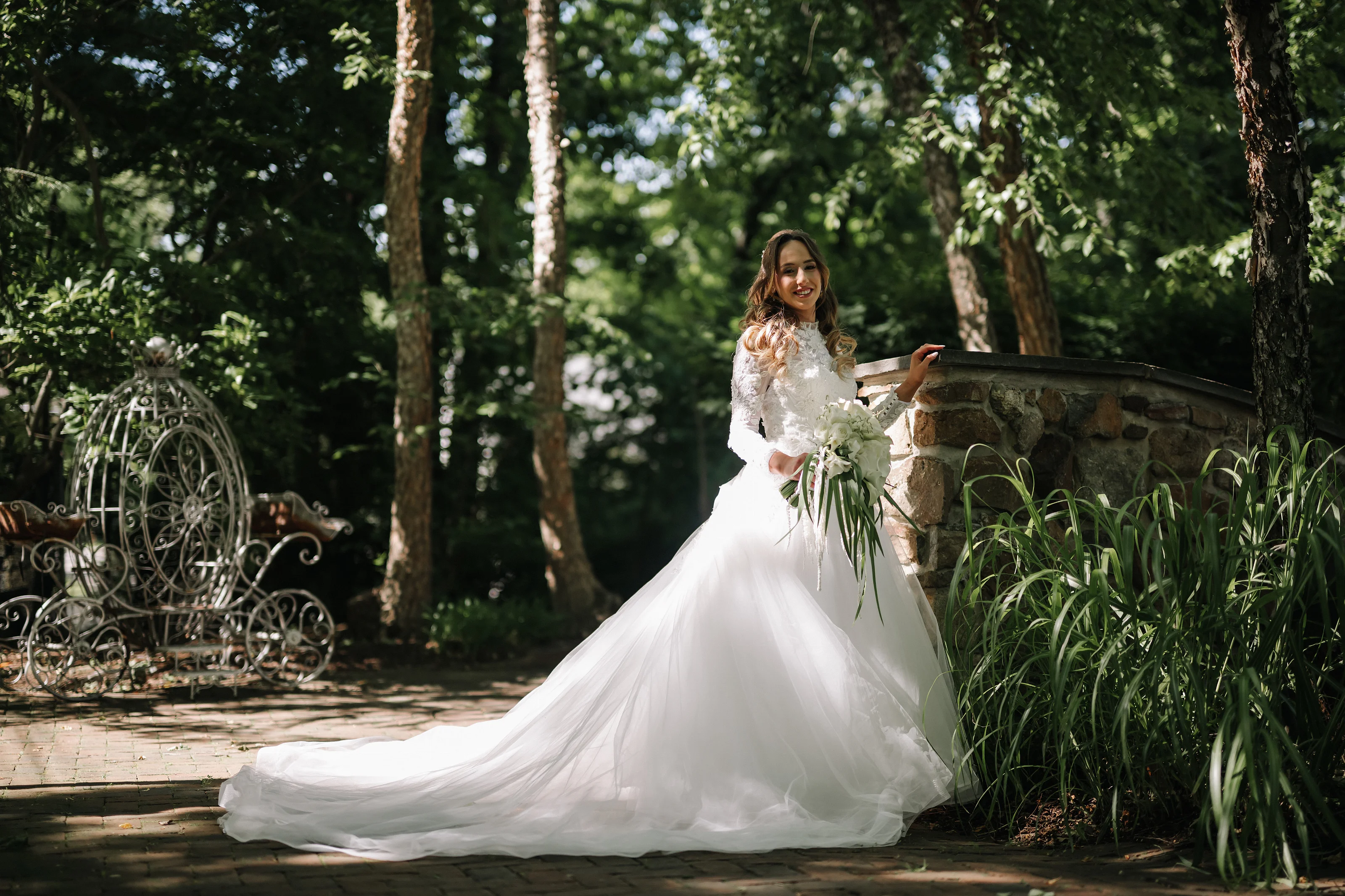 Bride smiling outdoors in a lush garden, holding her bouquet and lifting her wedding dress along a stone path.