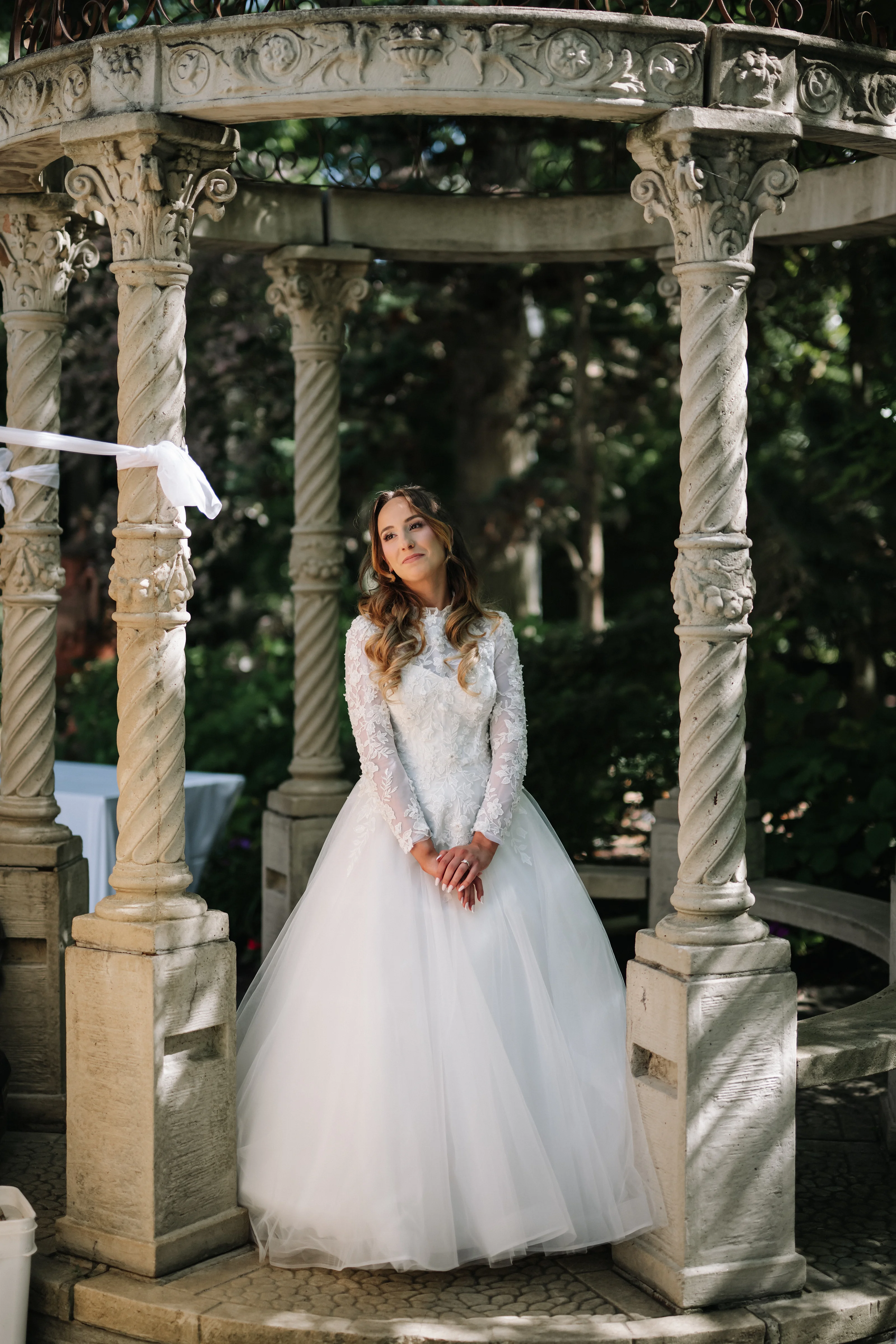 Bride in a lace long-sleeve wedding dress and veil posed under ornate stone arches in a garden, looking upward.