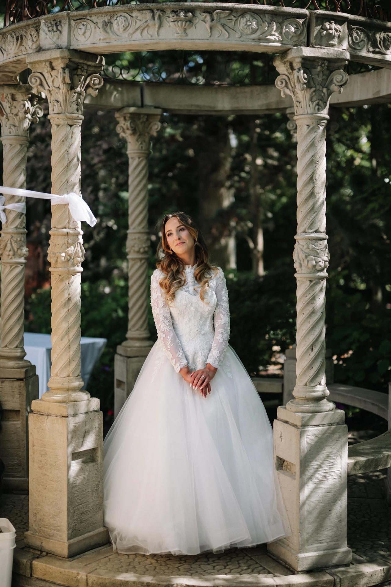 Bride in a lace long-sleeve wedding dress and veil posed under ornate stone arches in a garden, looking upward. Wedding at Crystal Plaza, Livingston, NJ.