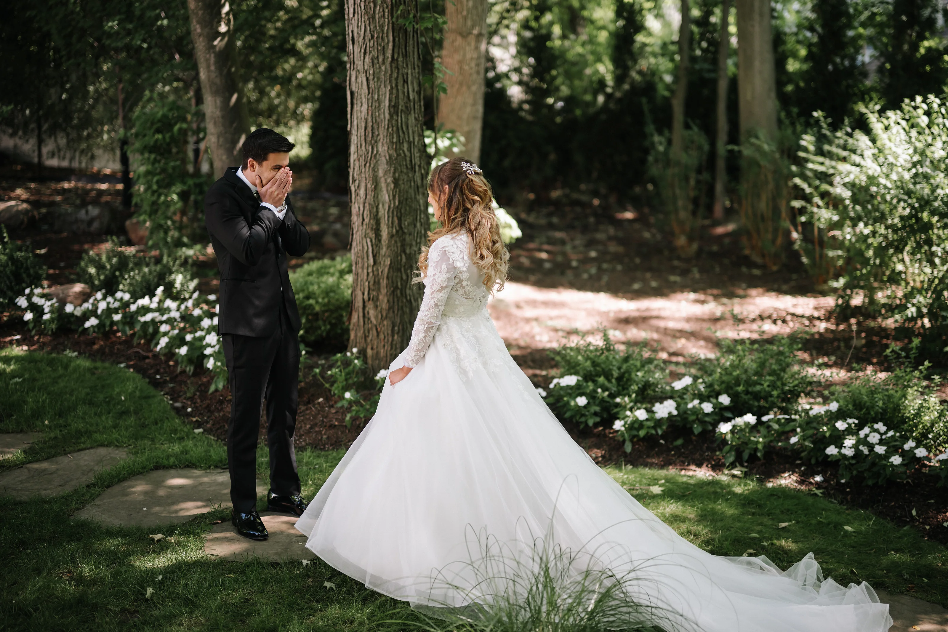 Groom covers his face with his hands as the bride in a lace gown walks toward him in a sunlit garden at their ceremony.