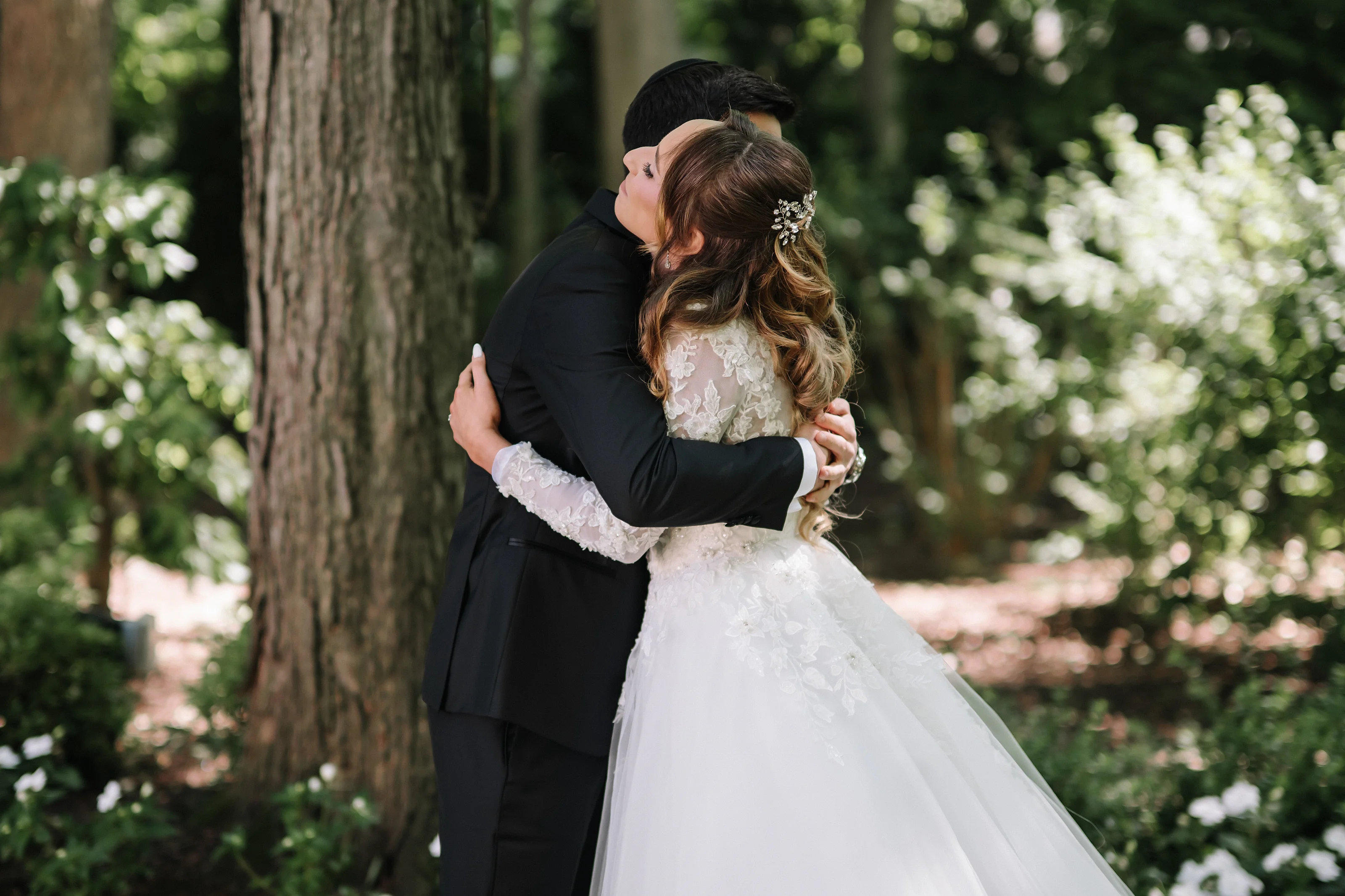 Bride and groom embracing closely outdoors, foreheads touching, with greenery and trees softly blurred in the background.