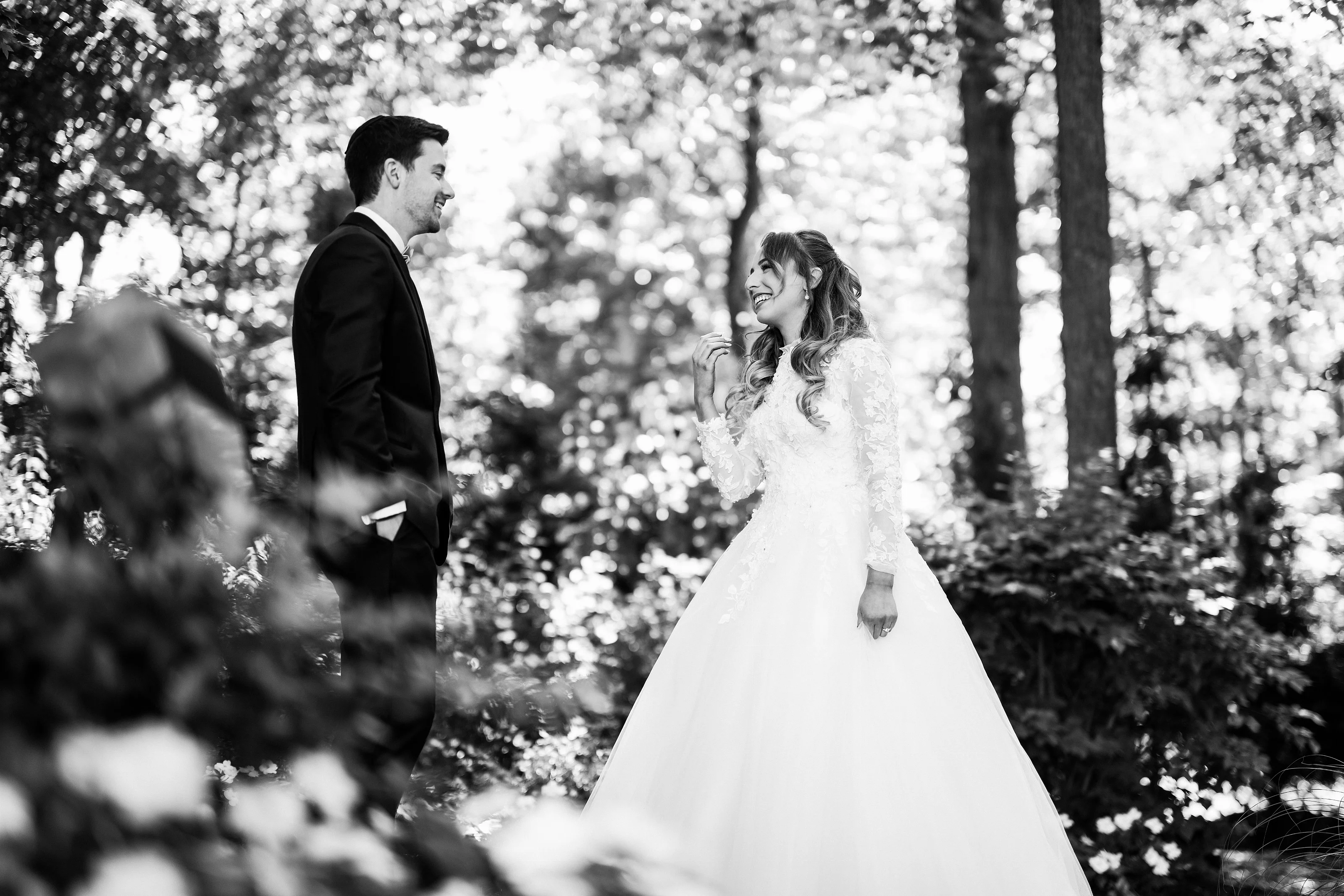 Black-and-white outdoor wedding photo of the bride and groom smiling at each other as confetti drifts through the sunlit air.
