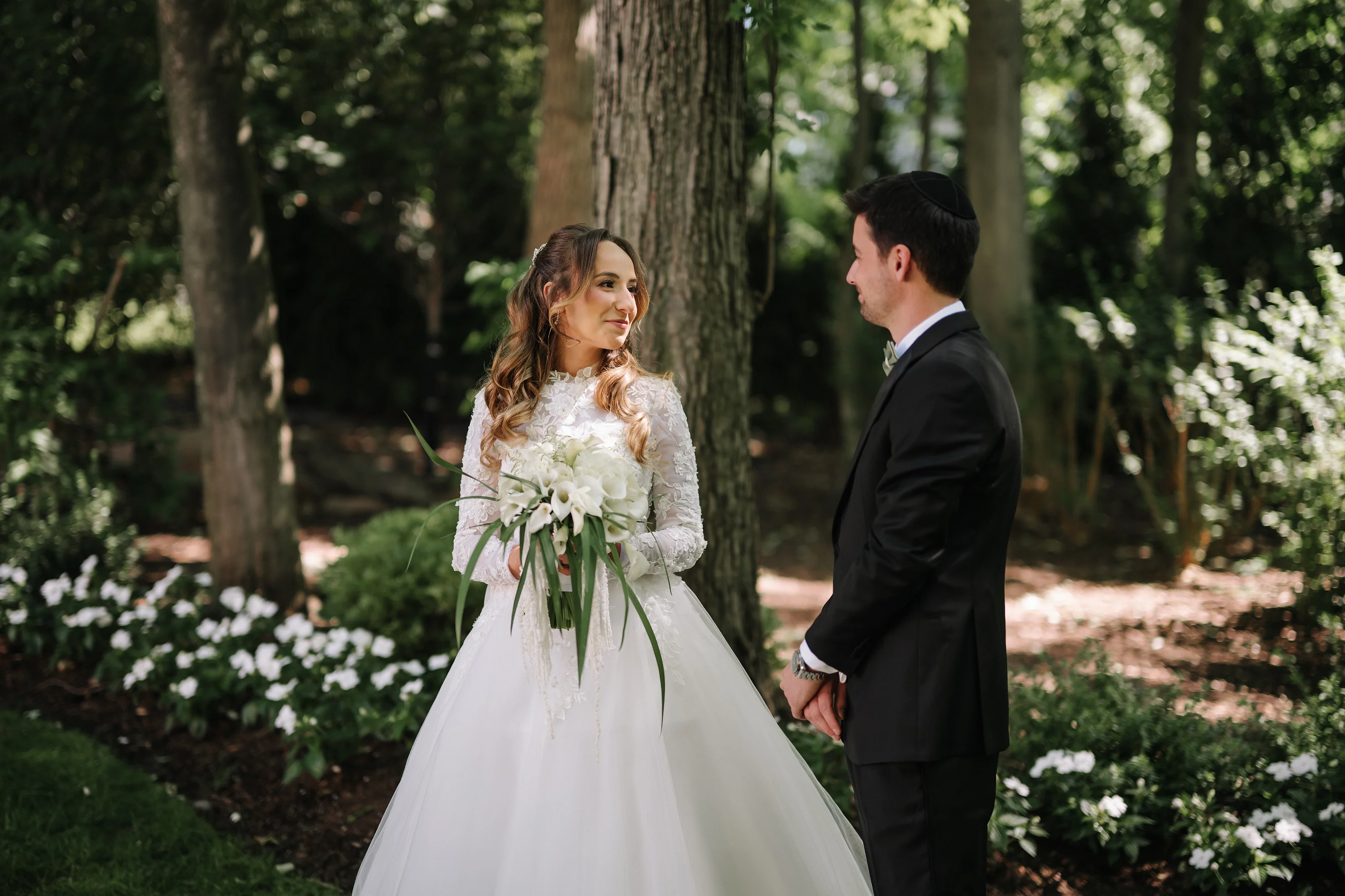Bride in a lace gown holding a white bouquet faces the groom in a garden as they stand close under dappled trees.