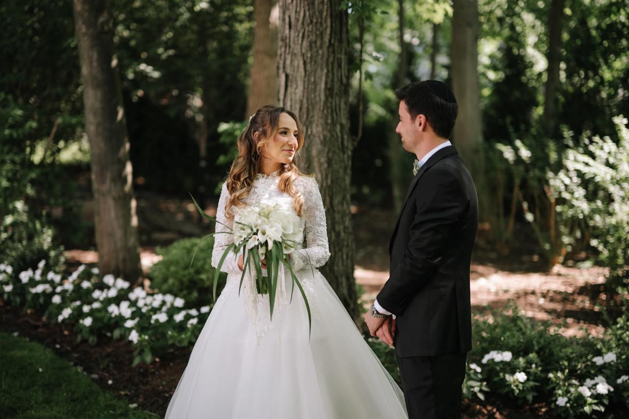 Bride in a lace gown holding a white bouquet faces the groom in a garden as they stand close under dappled trees. Wedding at Crystal Plaza, Livingston, NJ.