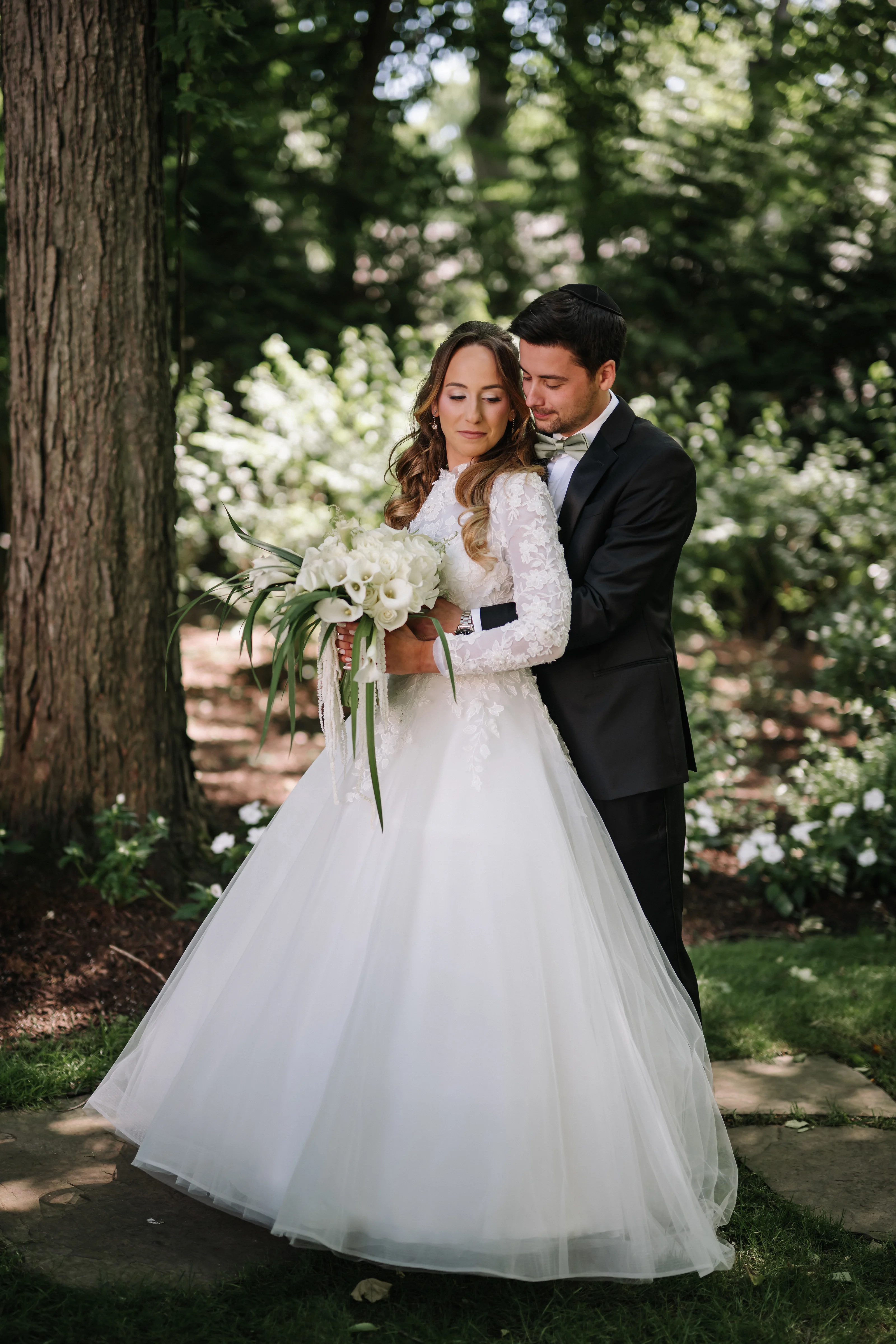 Bride and groom in a garden, embracing closely while the bride holds a bouquet of white flowers in natural light.