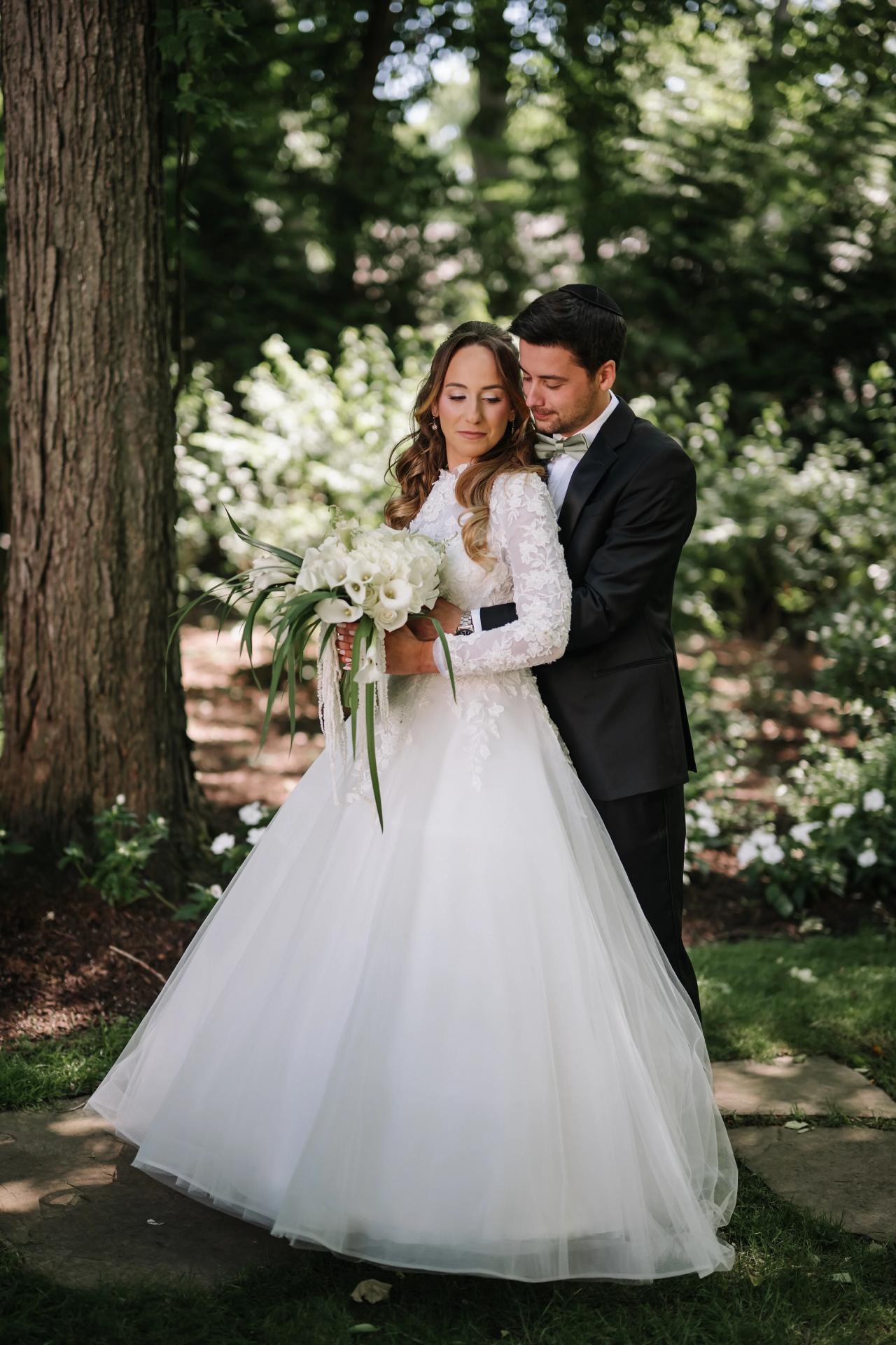 Bride and groom in a garden, embracing closely while the bride holds a bouquet of white flowers in natural light. Wedding at Crystal Plaza, Livingston, NJ.