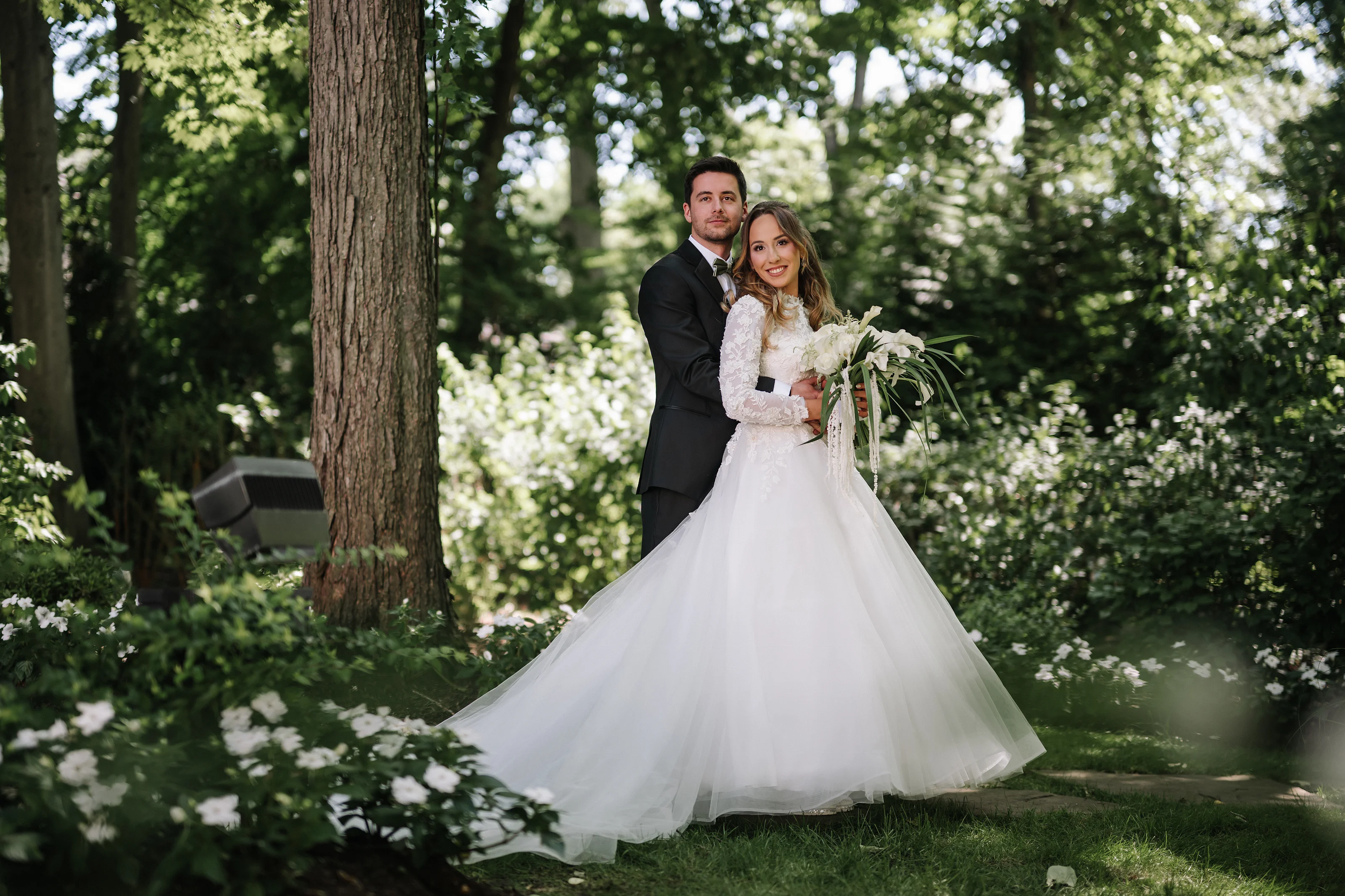 Wedding couple standing together outdoors in a garden, the bride holding a bouquet with a flowing veil while trees and greenery fill the background.