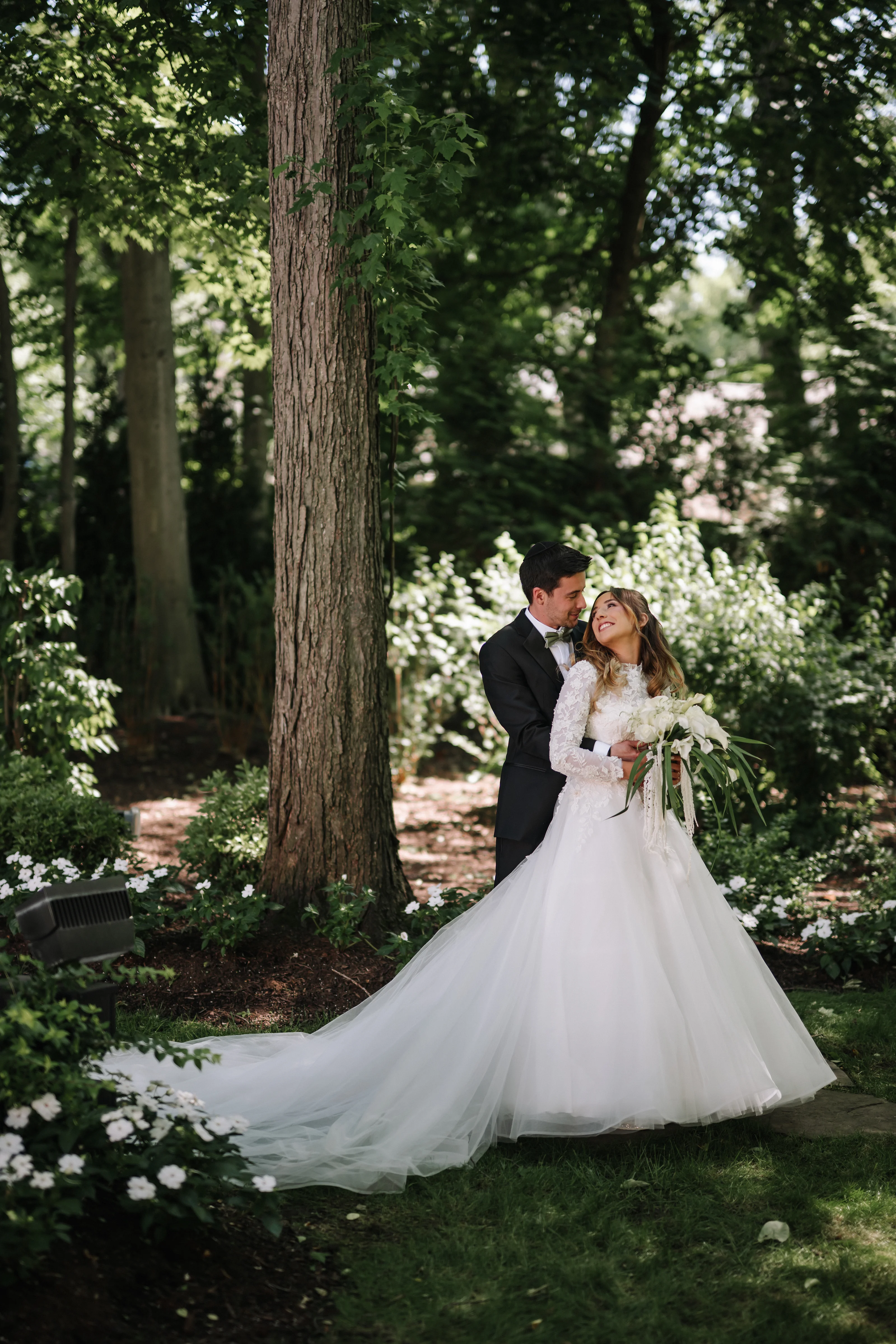 Newlyweds embracing in a lush garden with tall trees and soft natural light filtering through foliage.