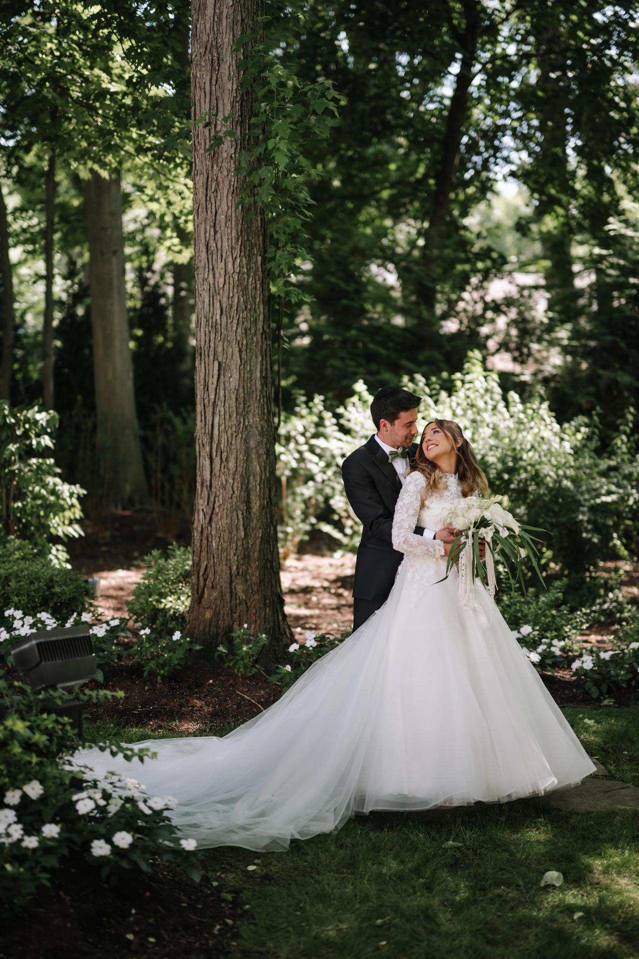 Newlyweds embracing in a lush garden with tall trees and soft natural light filtering through foliage. Wedding at Crystal Plaza, Livingston, NJ.