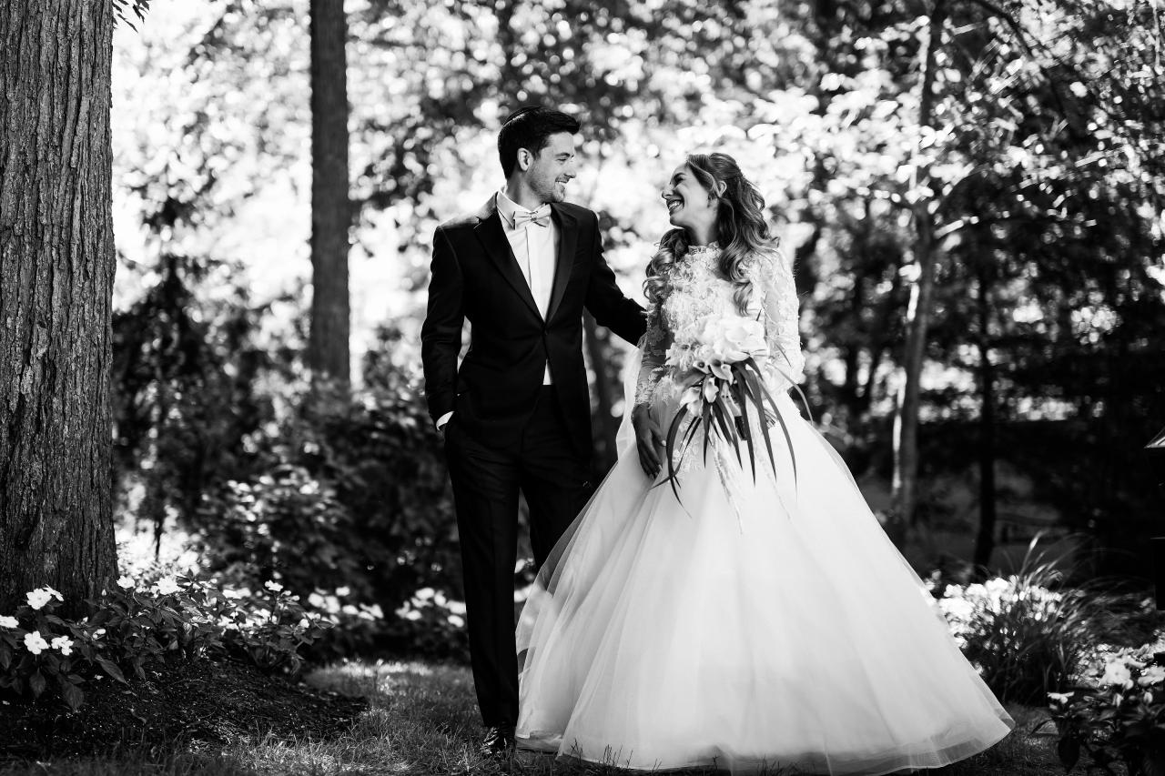 Black-and-white outdoor wedding portrait of a bride and groom walking hand in hand in dappled light through trees.