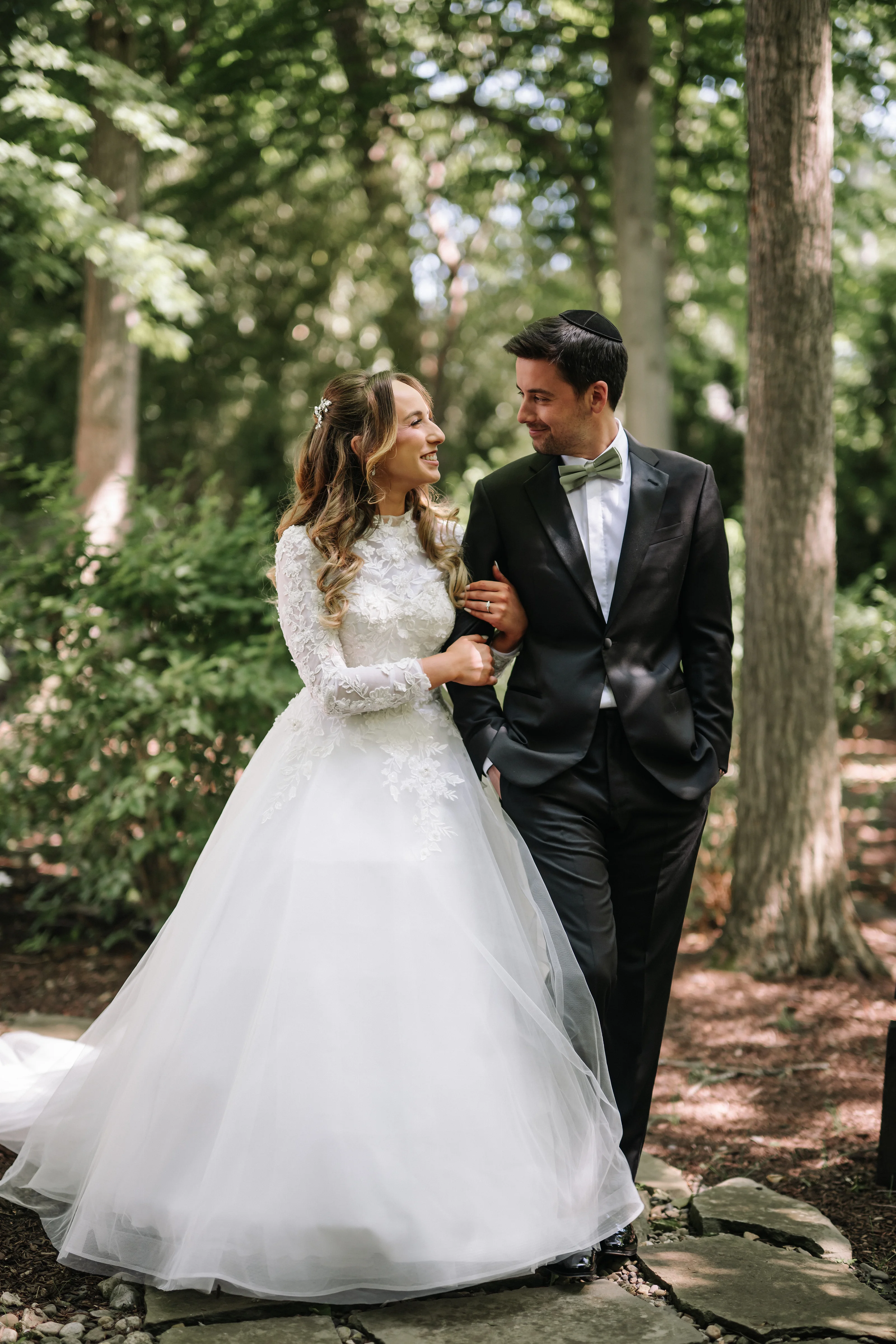 Bride and groom smiling at each other outdoors in a garden, holding hands as their wedding day portraits unfold among trees and greenery.