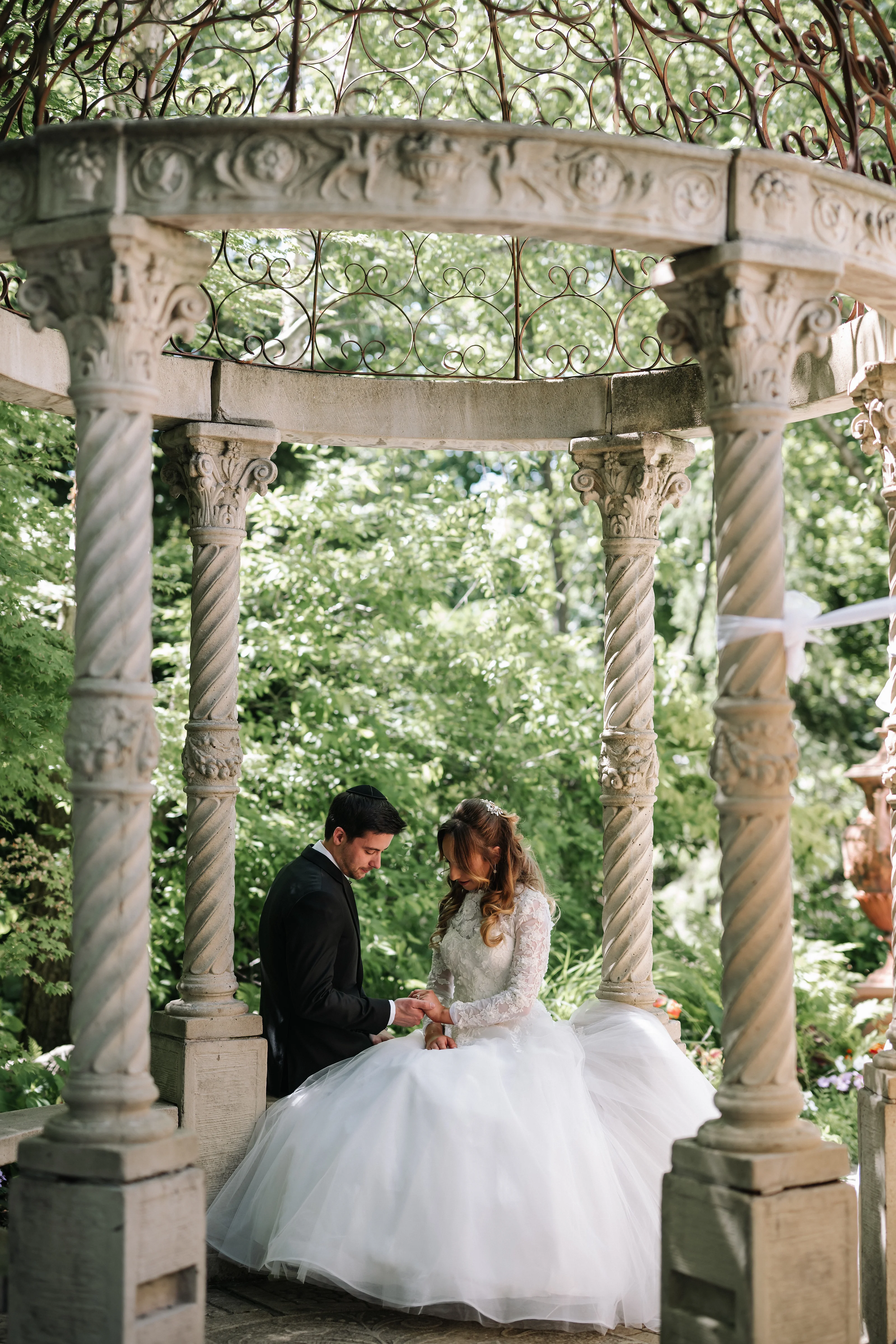 Ornate outdoor wedding scene with a bride and groom sharing an intimate moment under carved stone pillars and garden greenery.