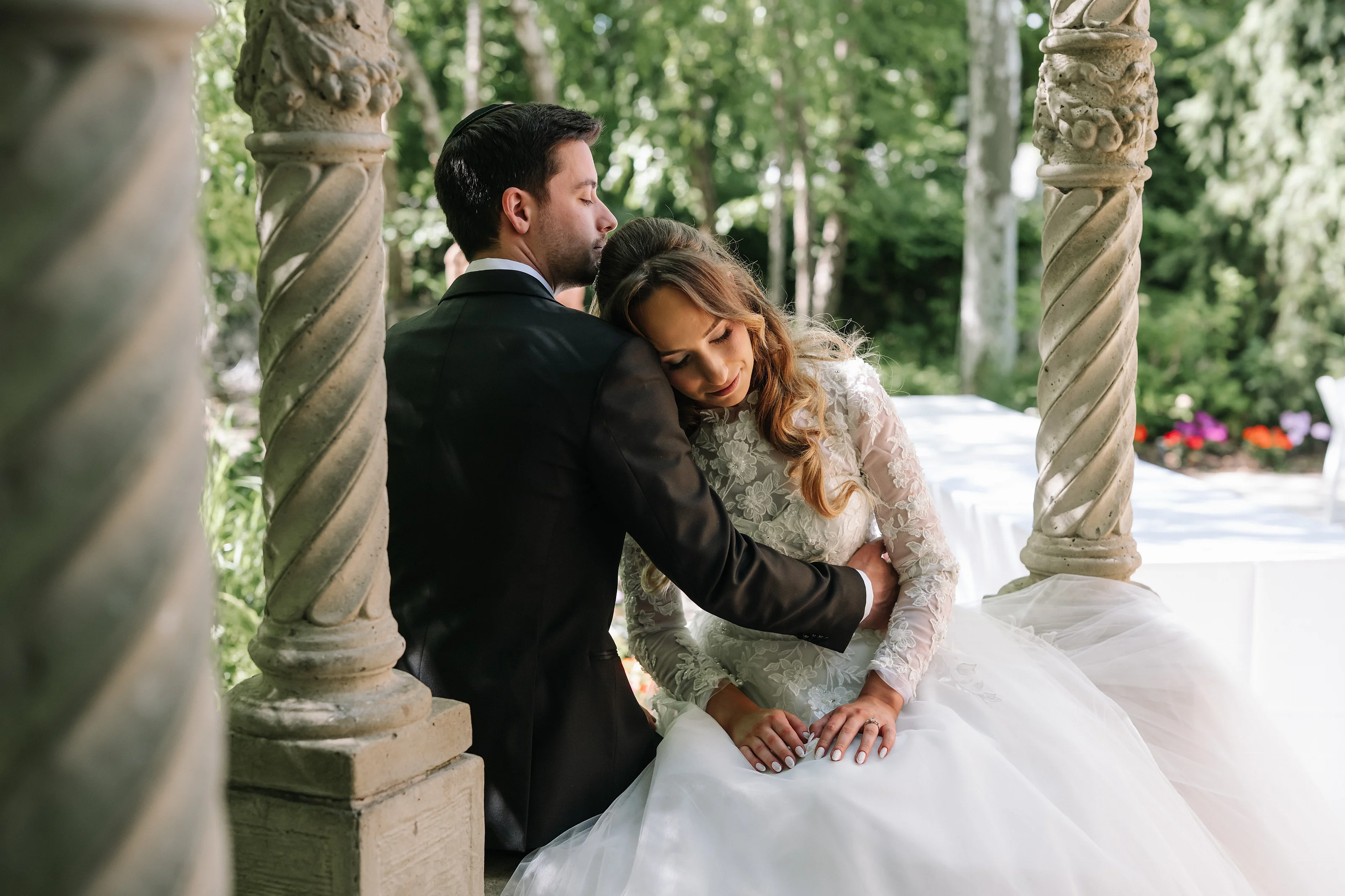 Wedding couple embracing closely outdoors beside stone columns, with the bride leaning into the groom during a quiet emotional moment.