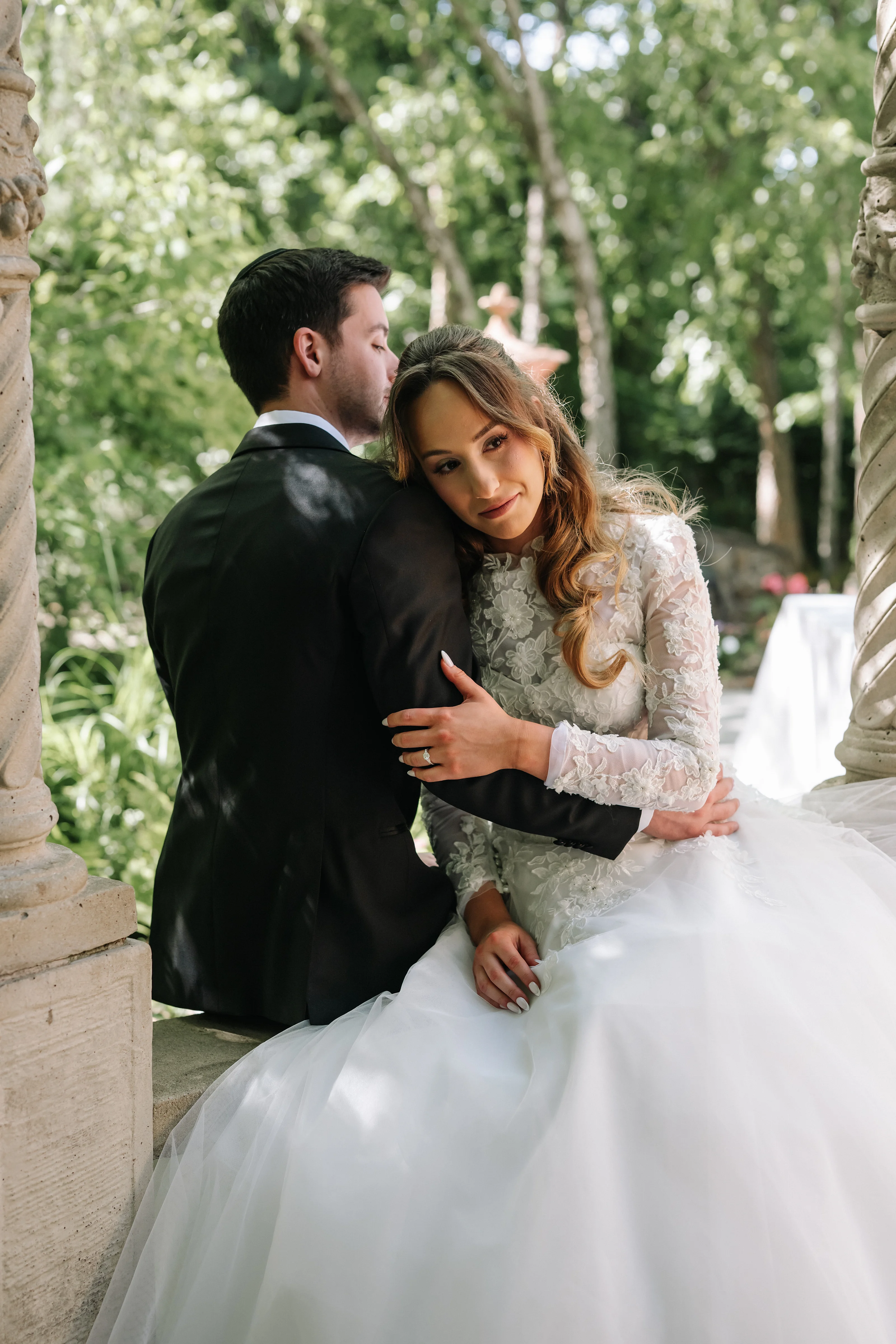 Bride and groom in a close embrace during outdoor wedding portraits, bride seated and smiling as groom leans in for a kiss, greenery background with soft bokeh.