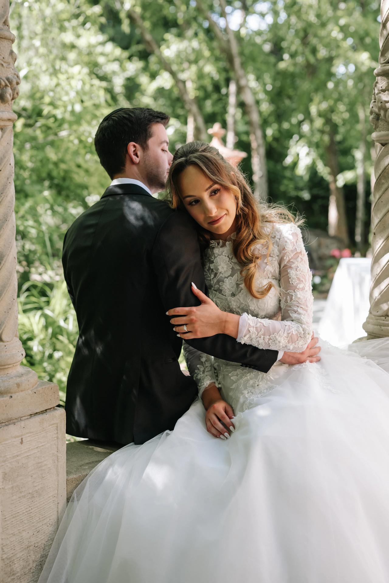 Bride and groom in a close embrace during outdoor wedding portraits, bride seated and smiling as groom leans in for a kiss, greenery background with soft bokeh. Wedding at Crystal Plaza, Livingston, NJ.