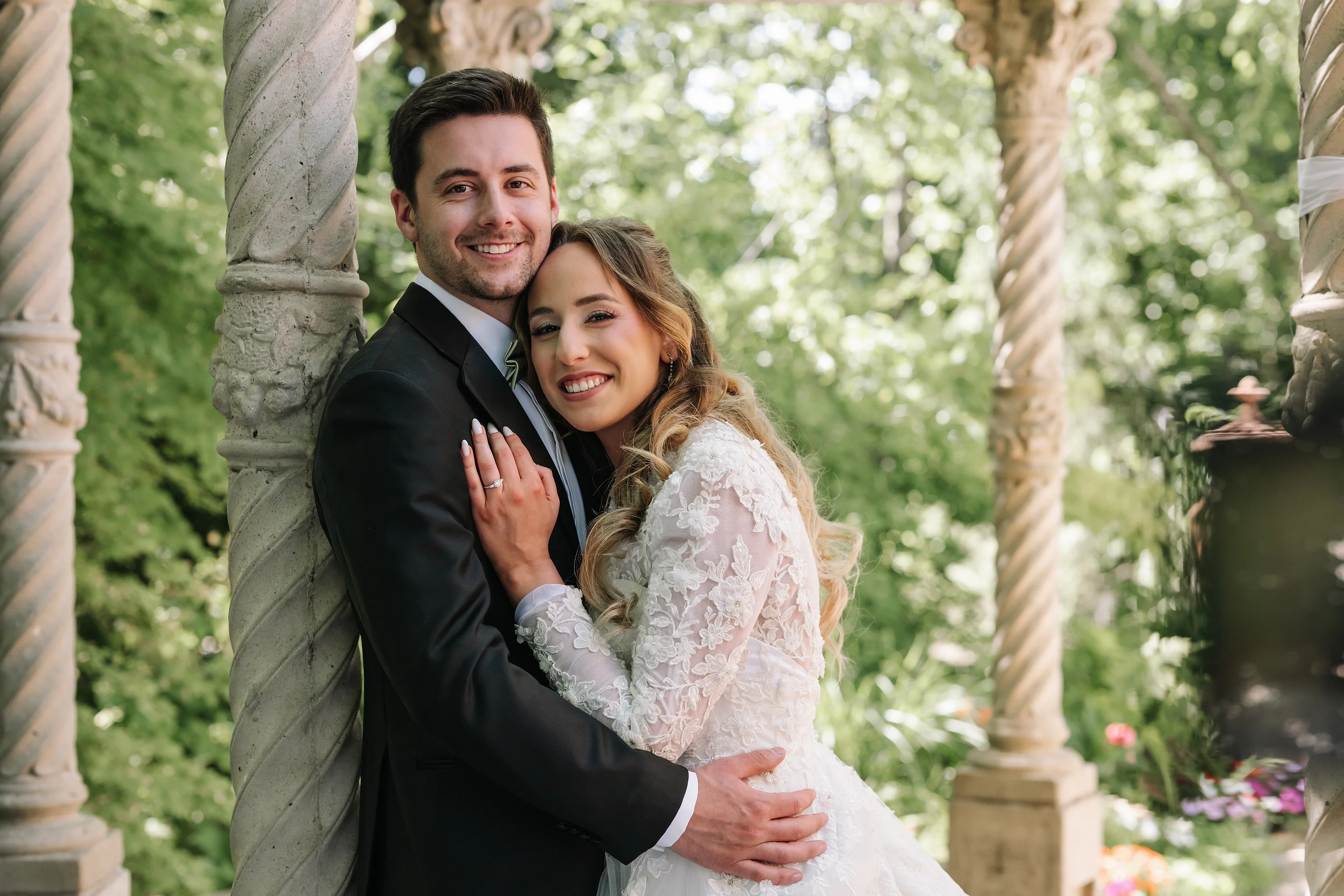 Bride and groom embracing and smiling during an outdoor wedding portrait with garden greenery and stone columns in the background.