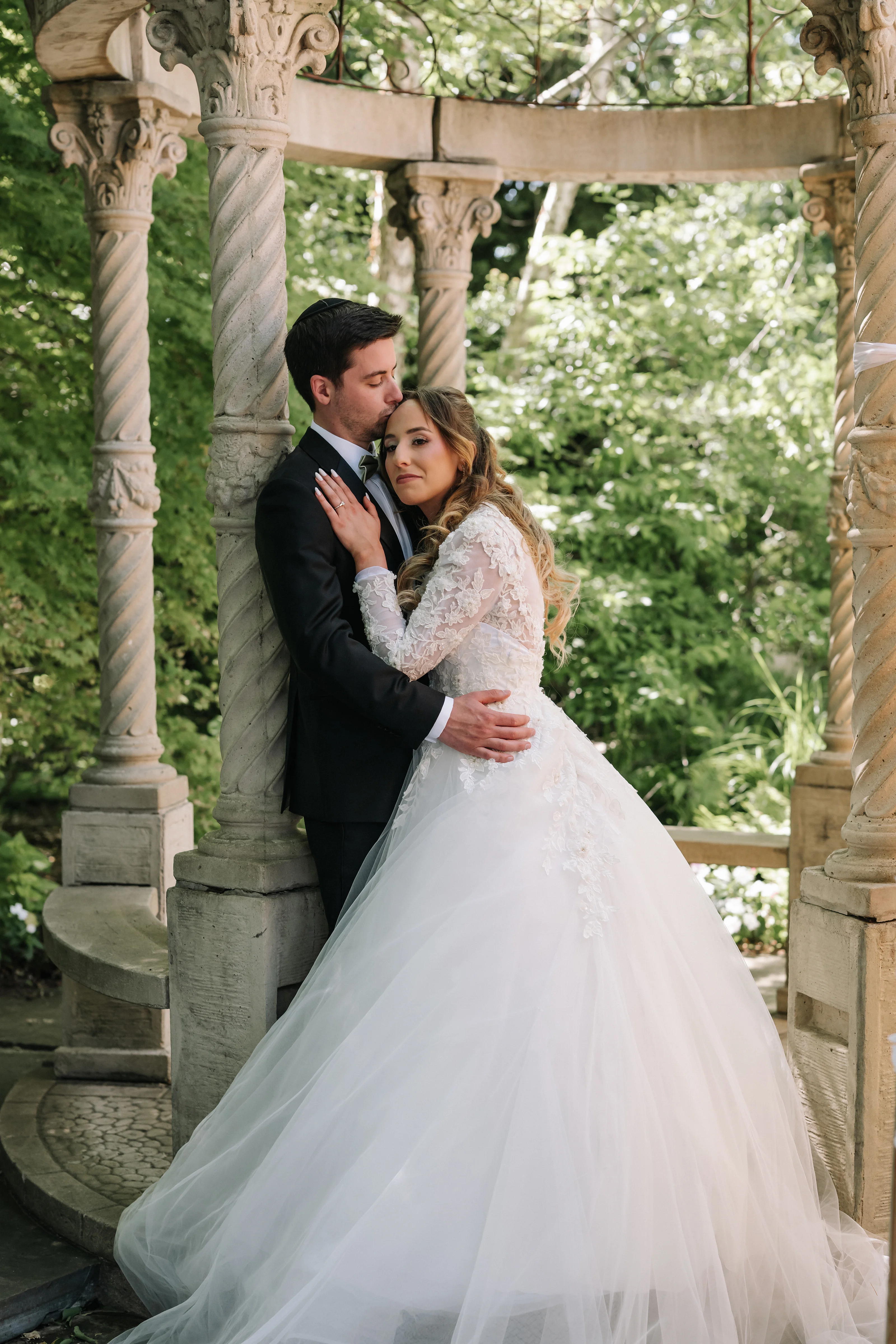 Bride and groom in a close embrace under a stone garden arch during badeken, soft daylight and greenery in the background.
