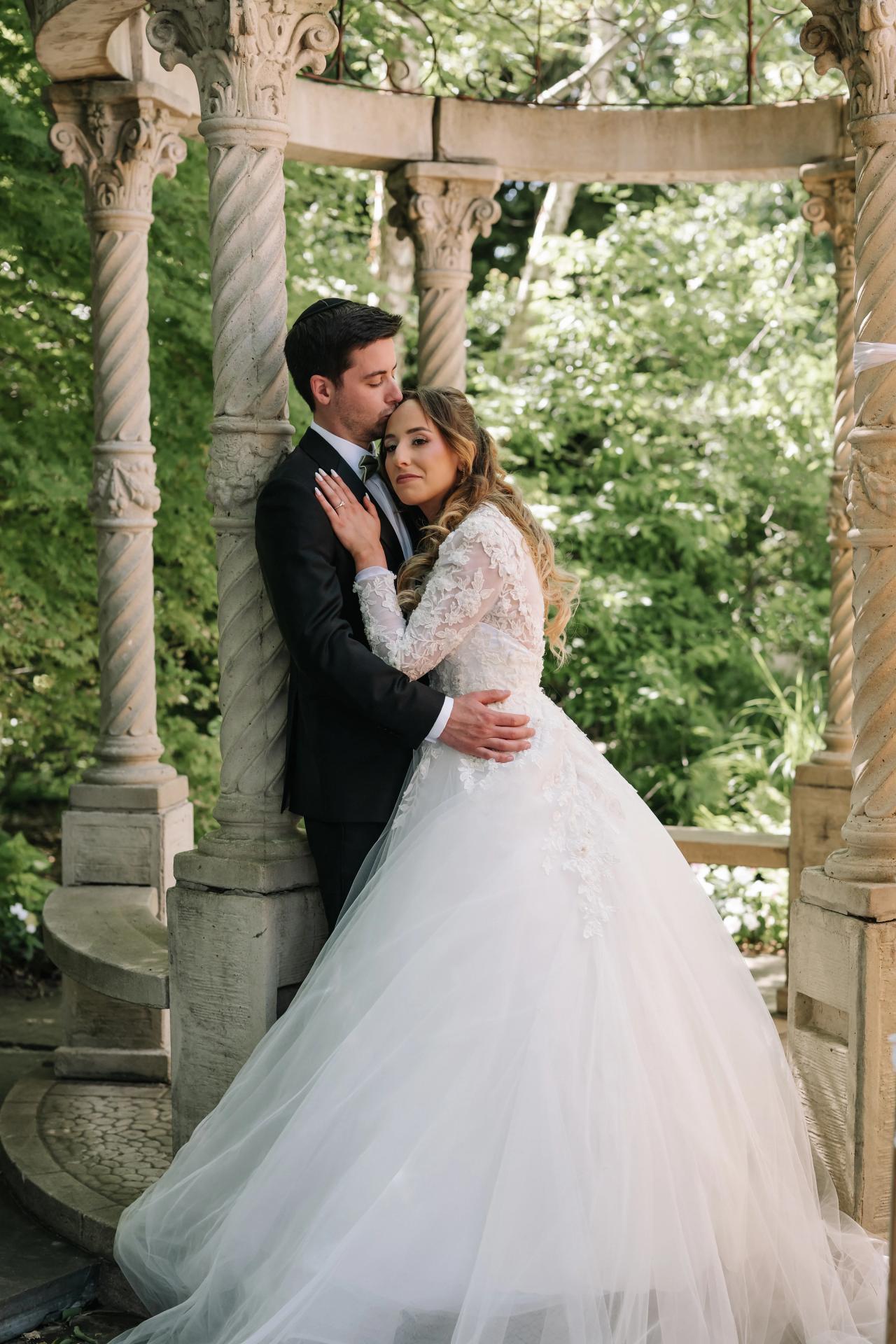 Bride and groom in a close embrace under a stone garden arch during badeken, soft daylight and greenery in the background. Wedding at Crystal Plaza, Livingston, NJ.