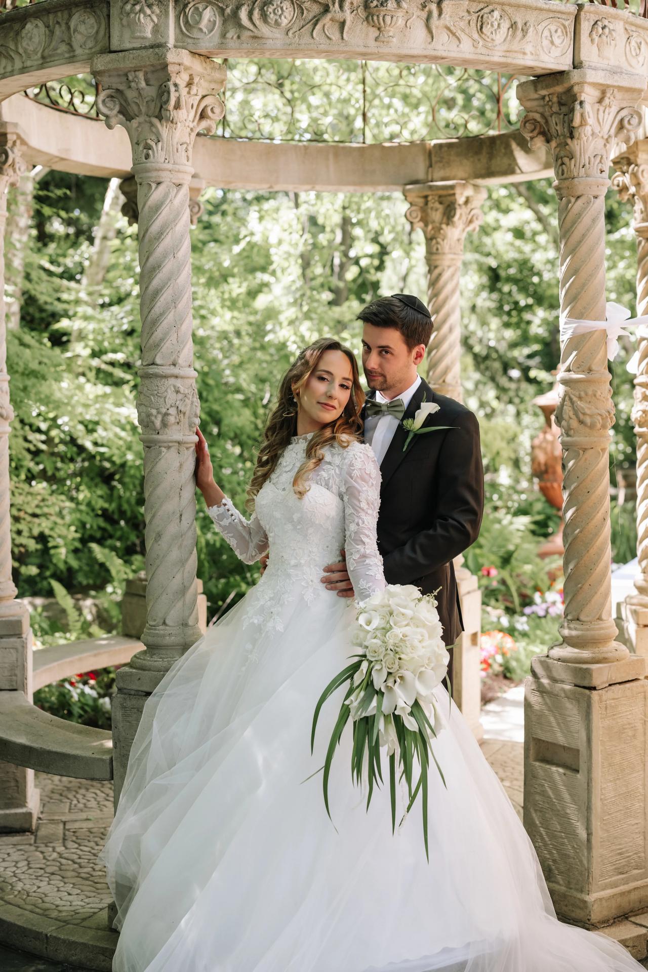 Wedding couple embracing and framed by ornate stone arches outdoors, bride in lace gown holding her veil and holding a bouquet, soft daylight and lush greenery behind.