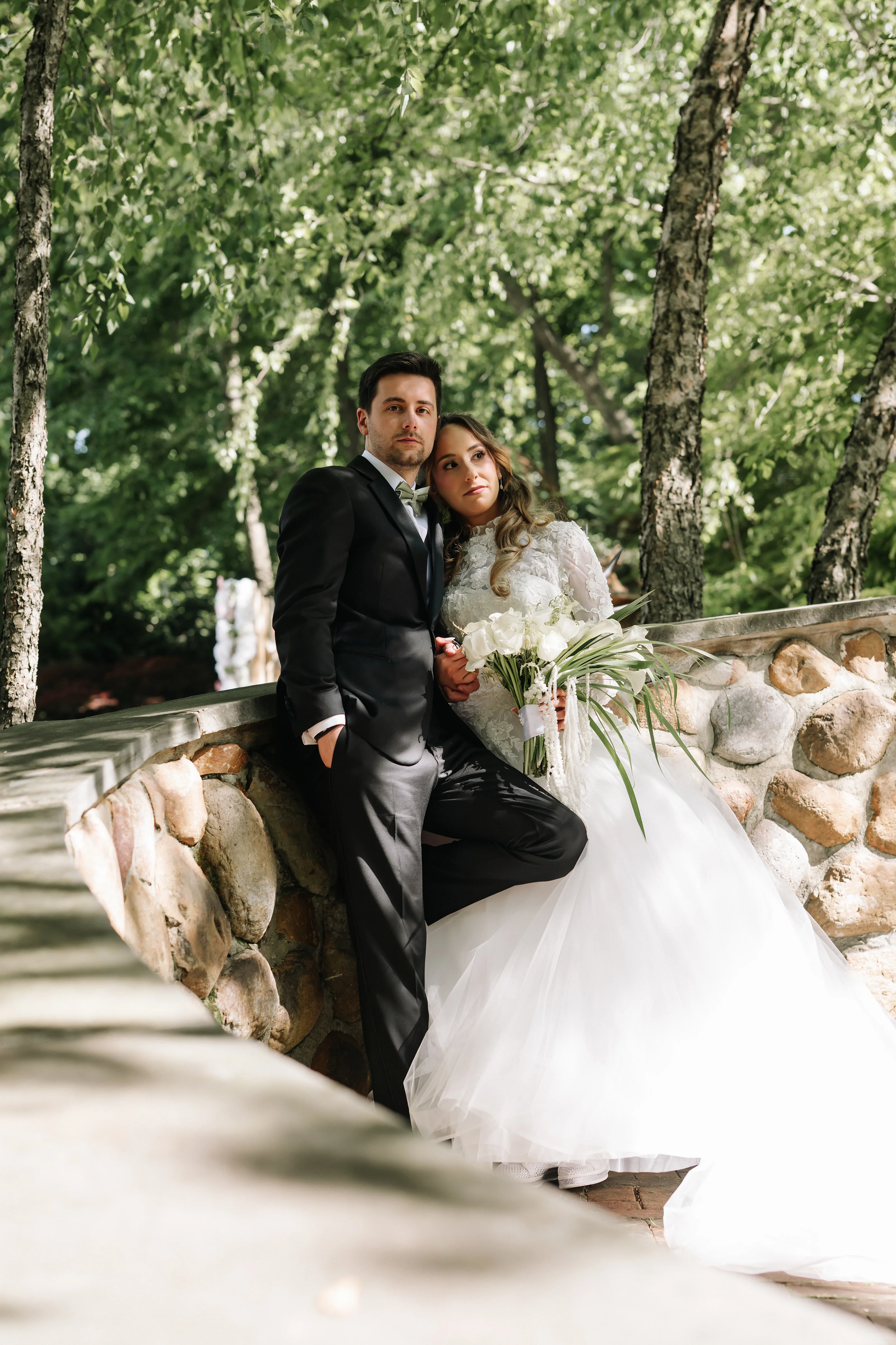 Bride and groom seated close together outdoors, bride holding a bouquet with veil flowing, framed by stone wall and greenery.