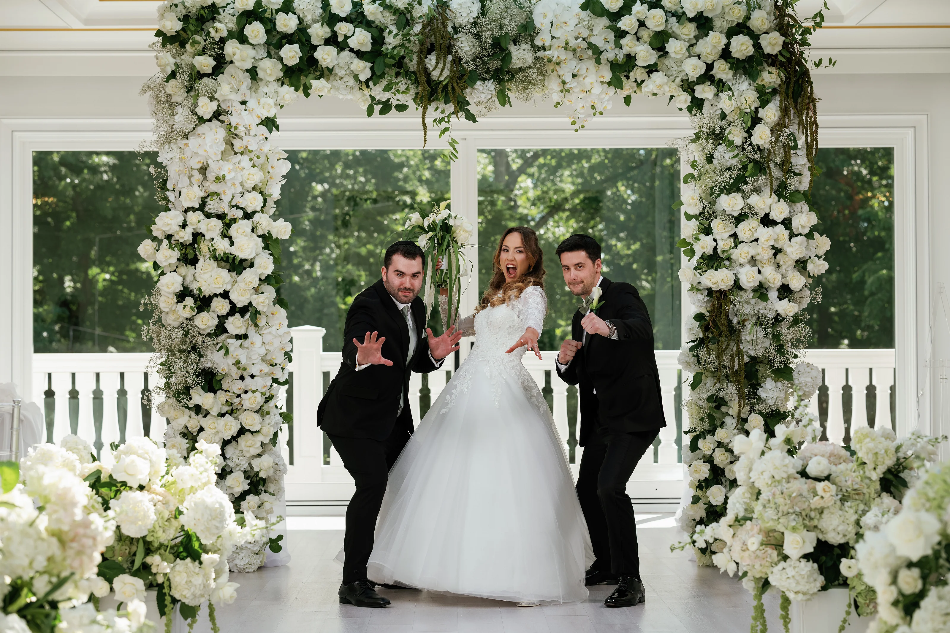 Bride laughing and walking forward at the ceremony entrance under a white floral chuppah, with the groom and a close family member reacting with joyful hand gestures.