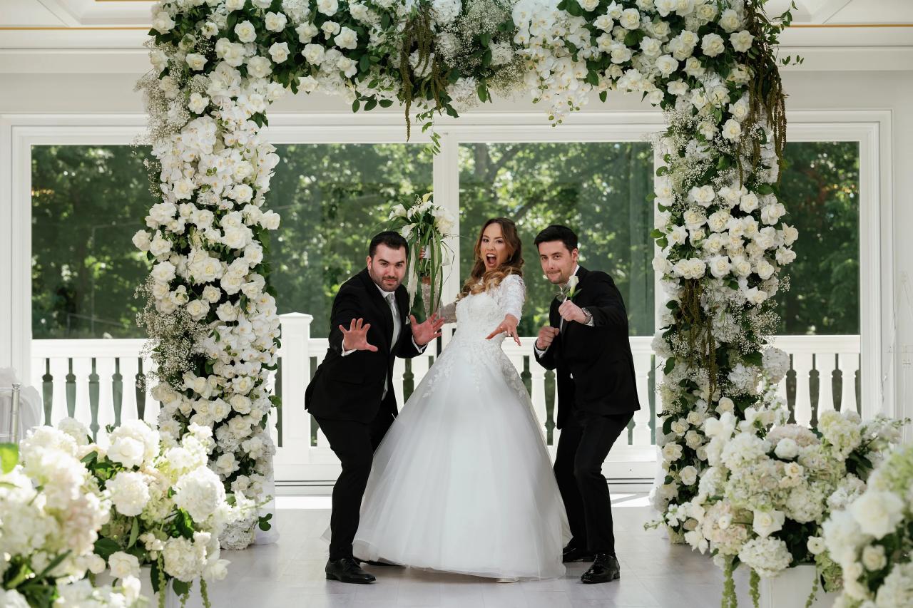 Bride laughing and walking forward at the ceremony entrance under a white floral chuppah, with the groom and a close family member reacting with joyful hand gestures. Wedding at Crystal Plaza, Livingston, NJ.