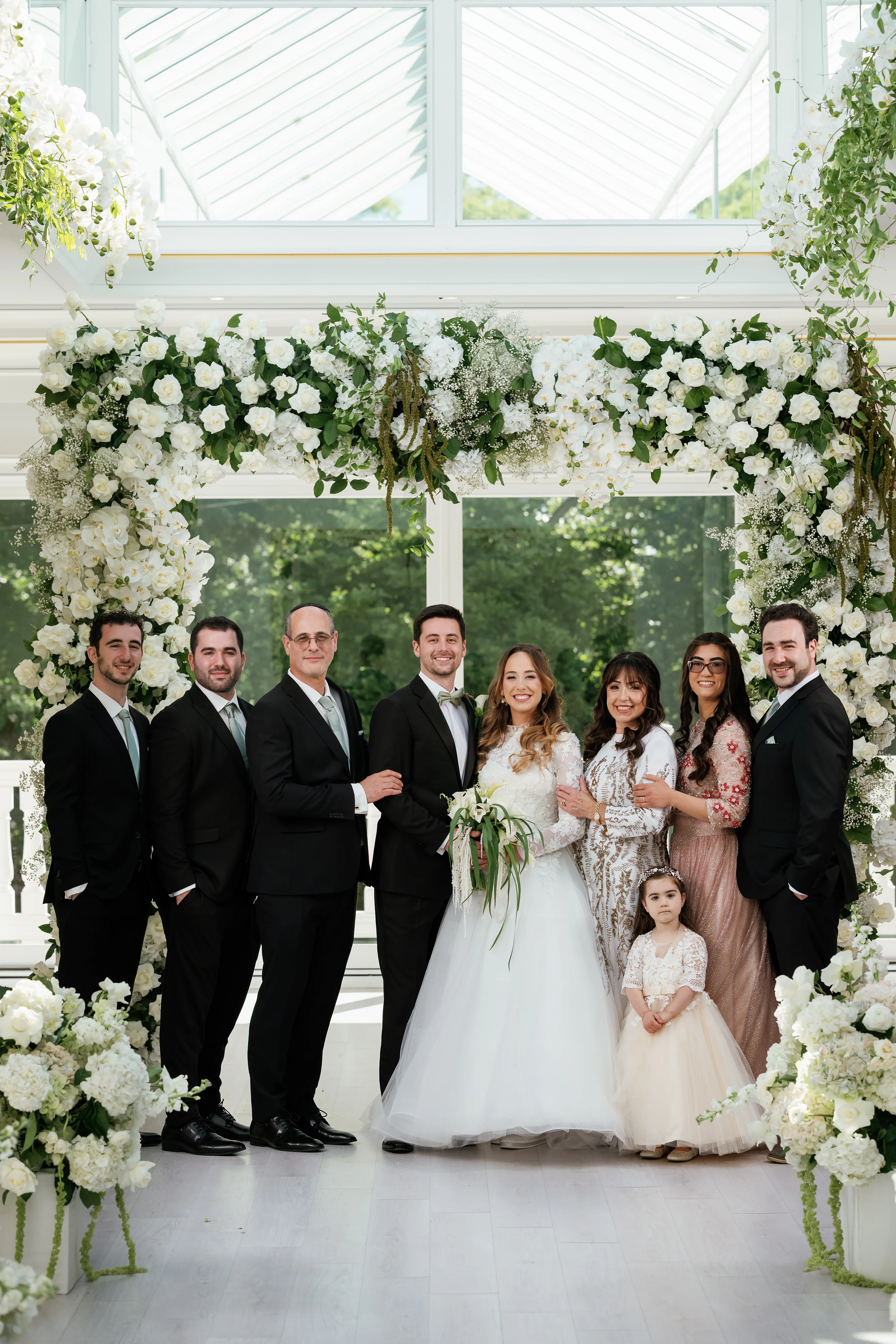 Wedding family formal group standing under a floral chuppah with the bride and groom centered, framed by white roses and greenery.