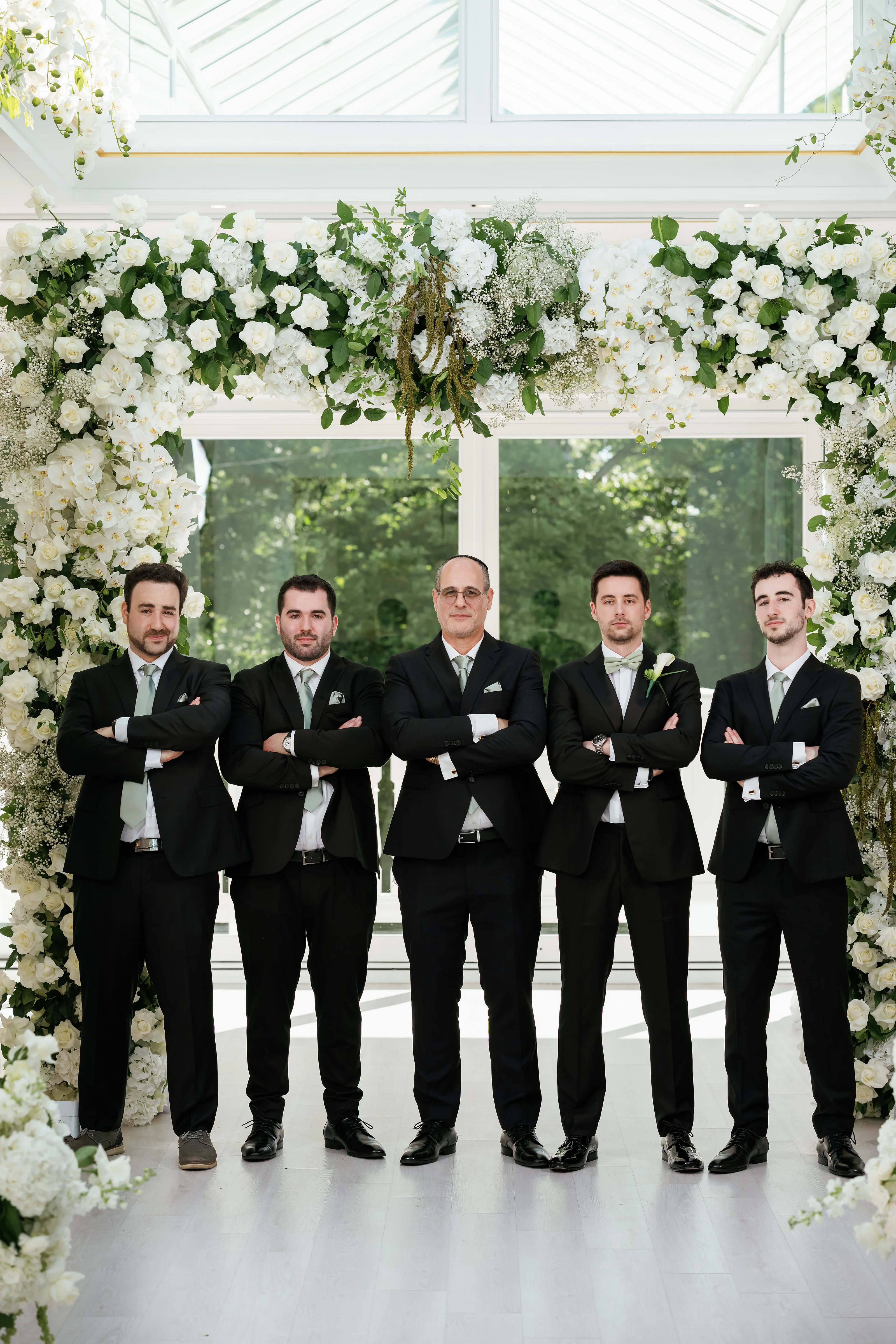 A formal group portrait of five men in black suits and bow ties standing beneath a floral wedding arch with white roses and greenery.