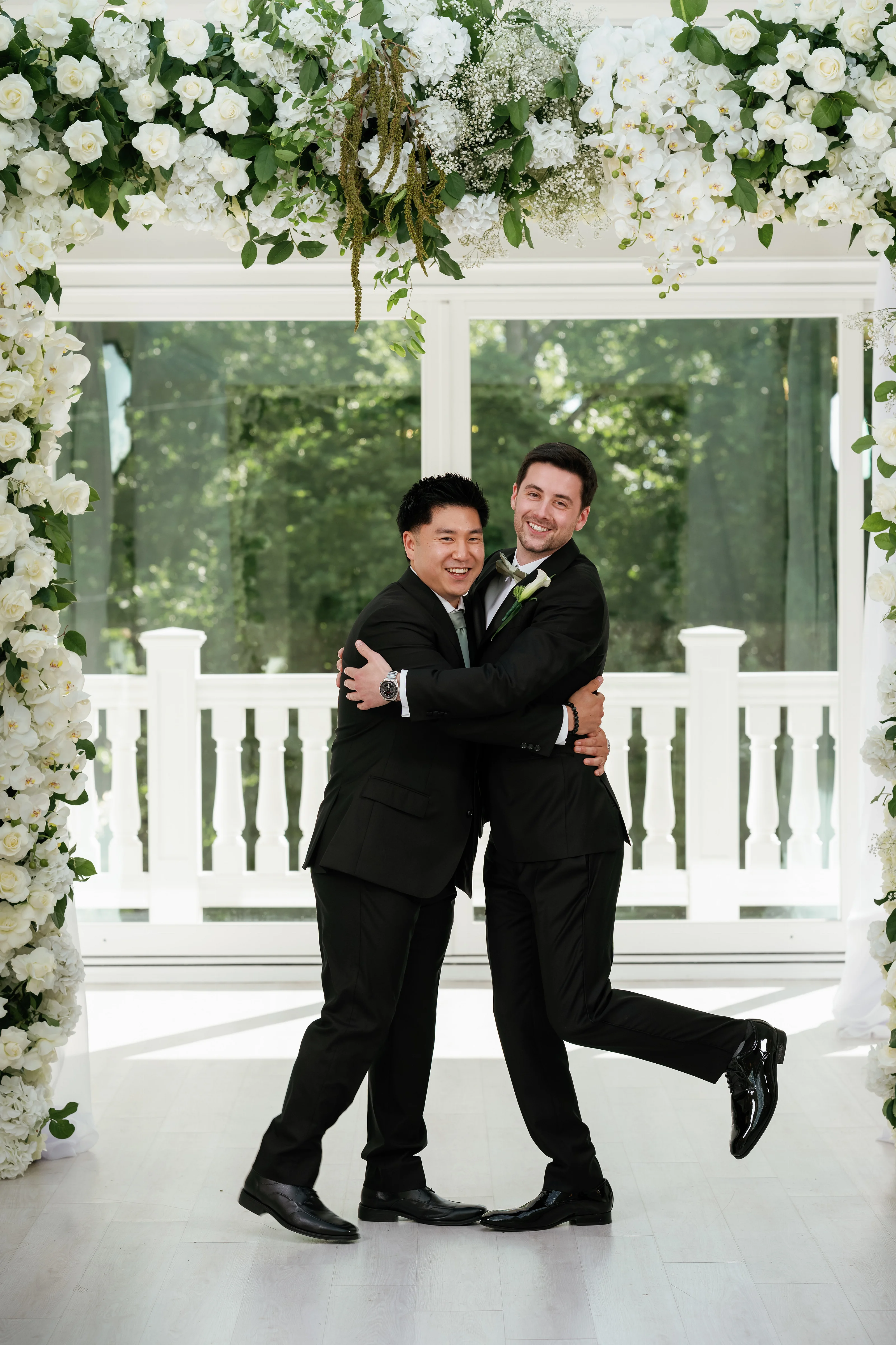 Smiling couple in formalwear embracing beneath a white floral arch with garden greenery in the background.