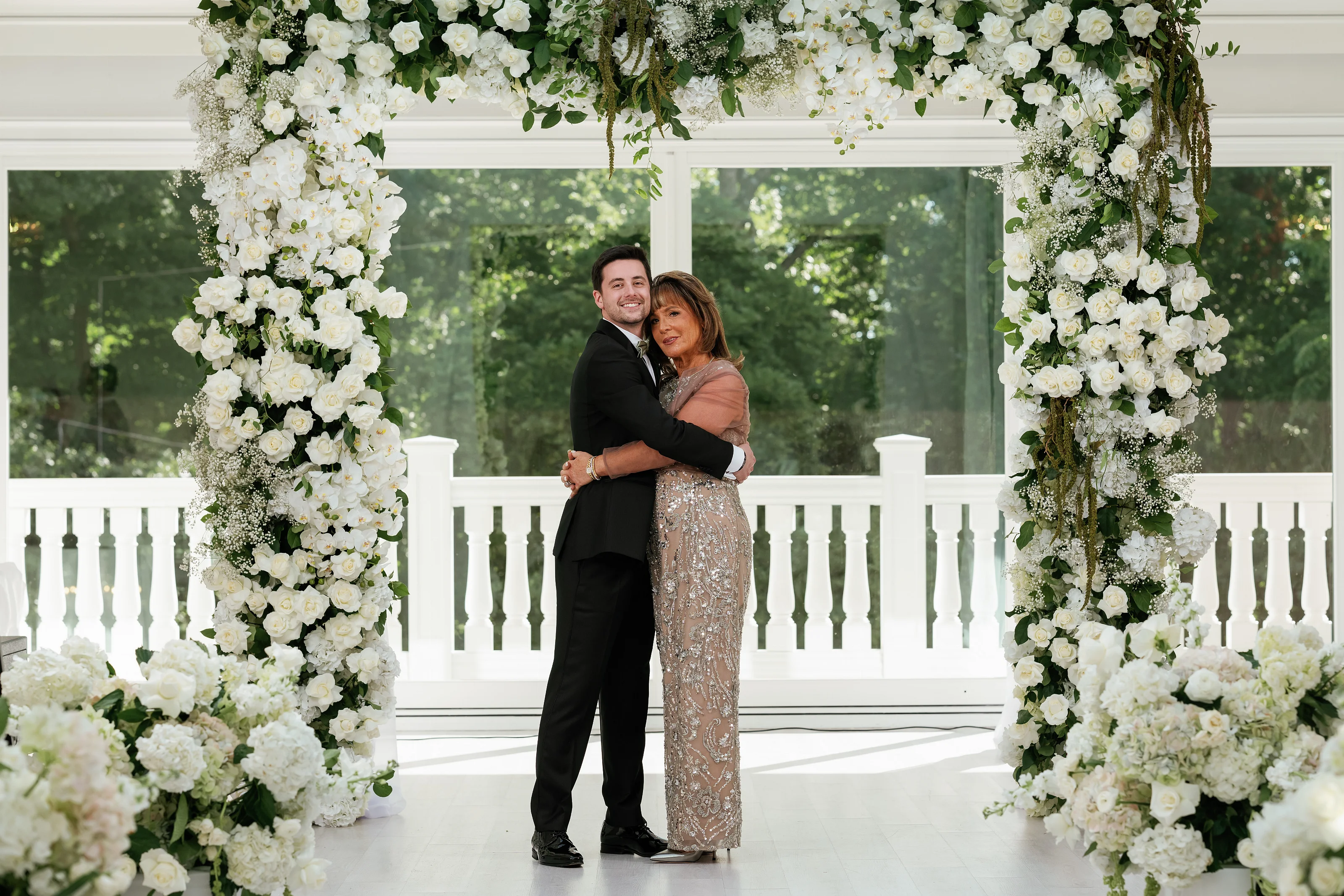 Wedding couple embracing under an elegant floral arch with abundant white flowers and greenery in bright daylight.