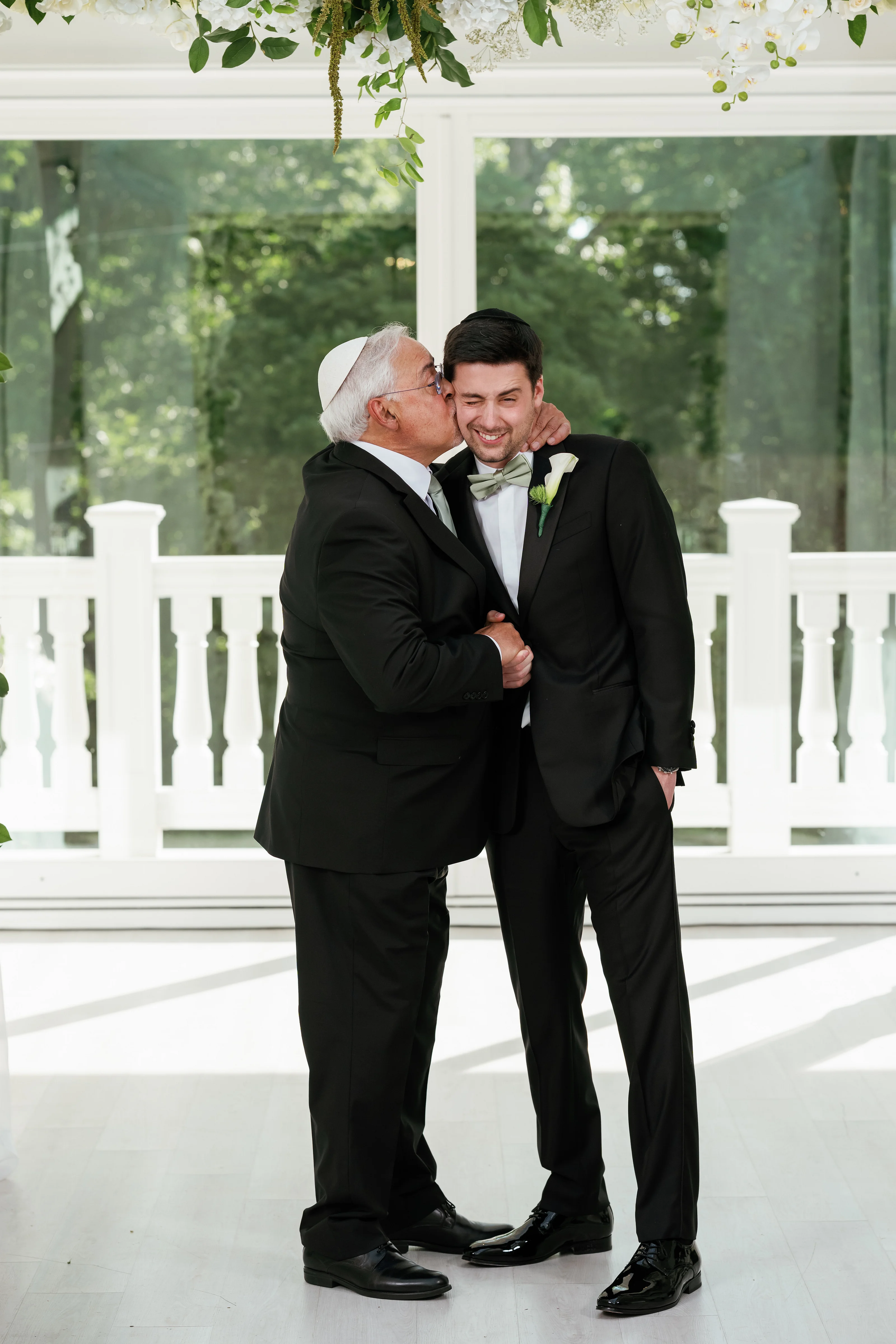 Bride’s badeken moment: groom in a black suit receiving a tender kiss and embrace as the elderly man leans in; framed by white railings and greenery.