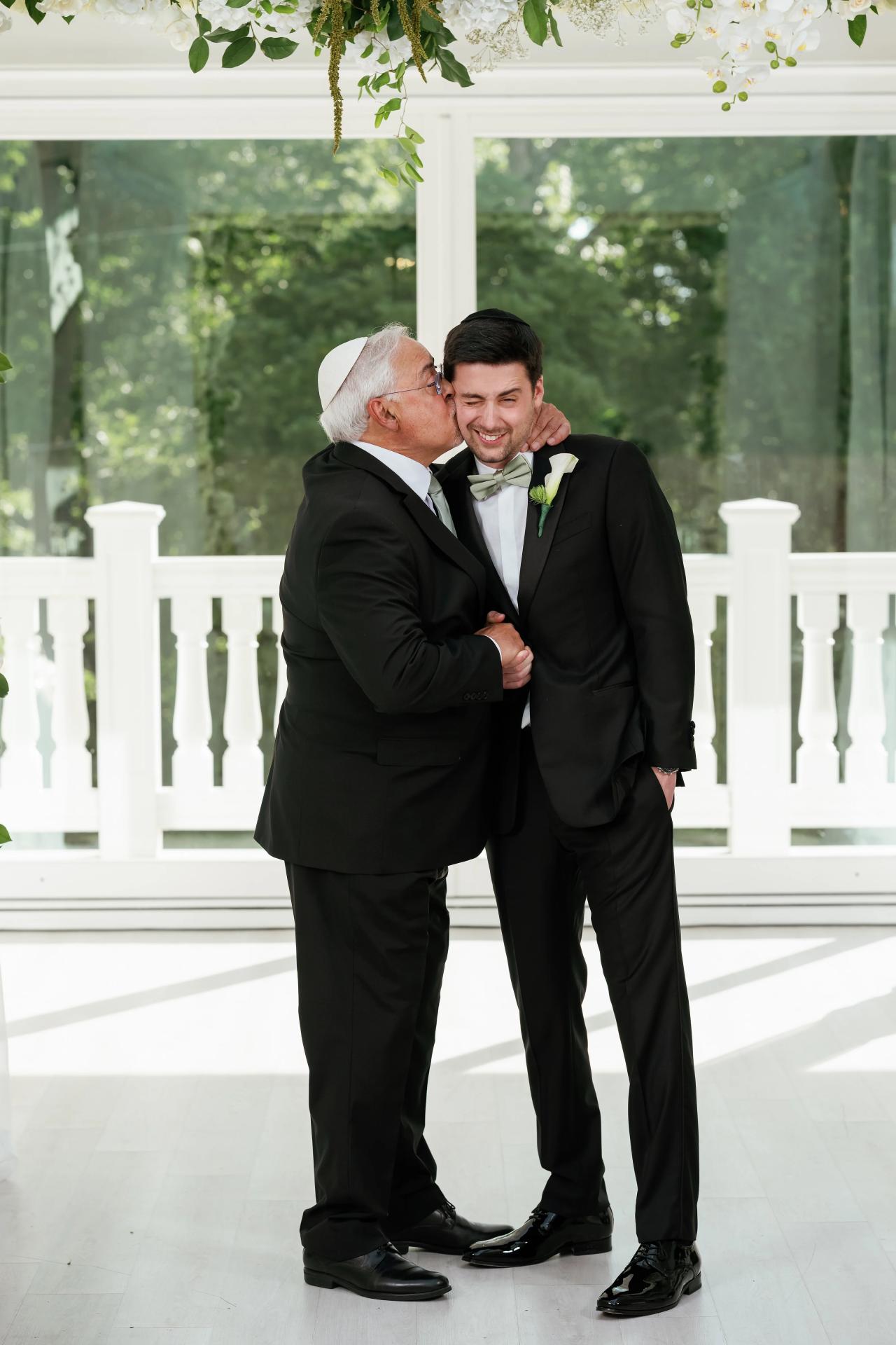 Bride’s badeken moment: groom in a black suit receiving a tender kiss and embrace as the elderly man leans in; framed by white railings and greenery. Wedding at Crystal Plaza, Livingston, NJ.