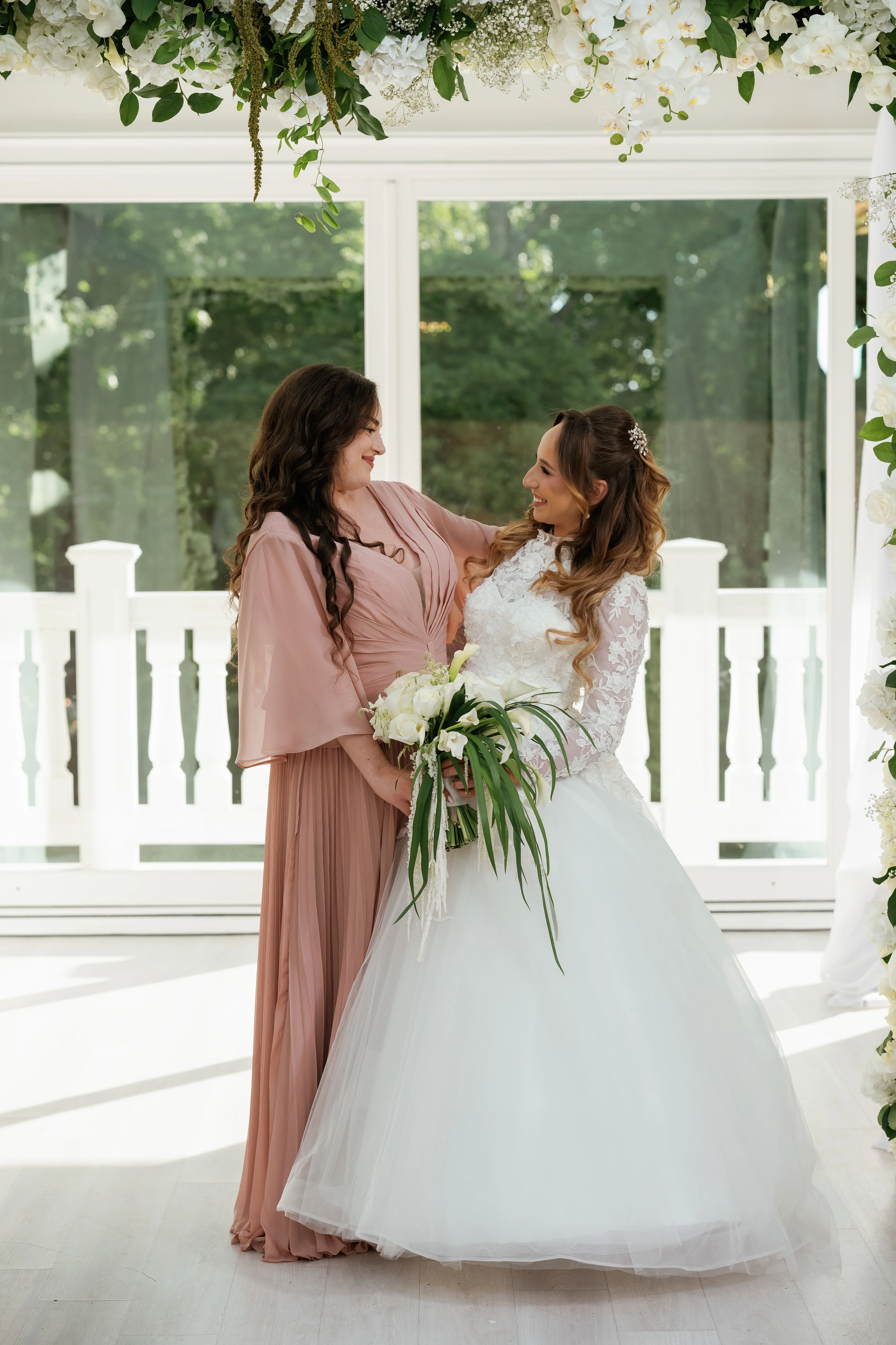 Bride in a white gown and veil sharing a tender embrace with a woman in a blush dress during the badeken moment, framed by white railing and floral decor.