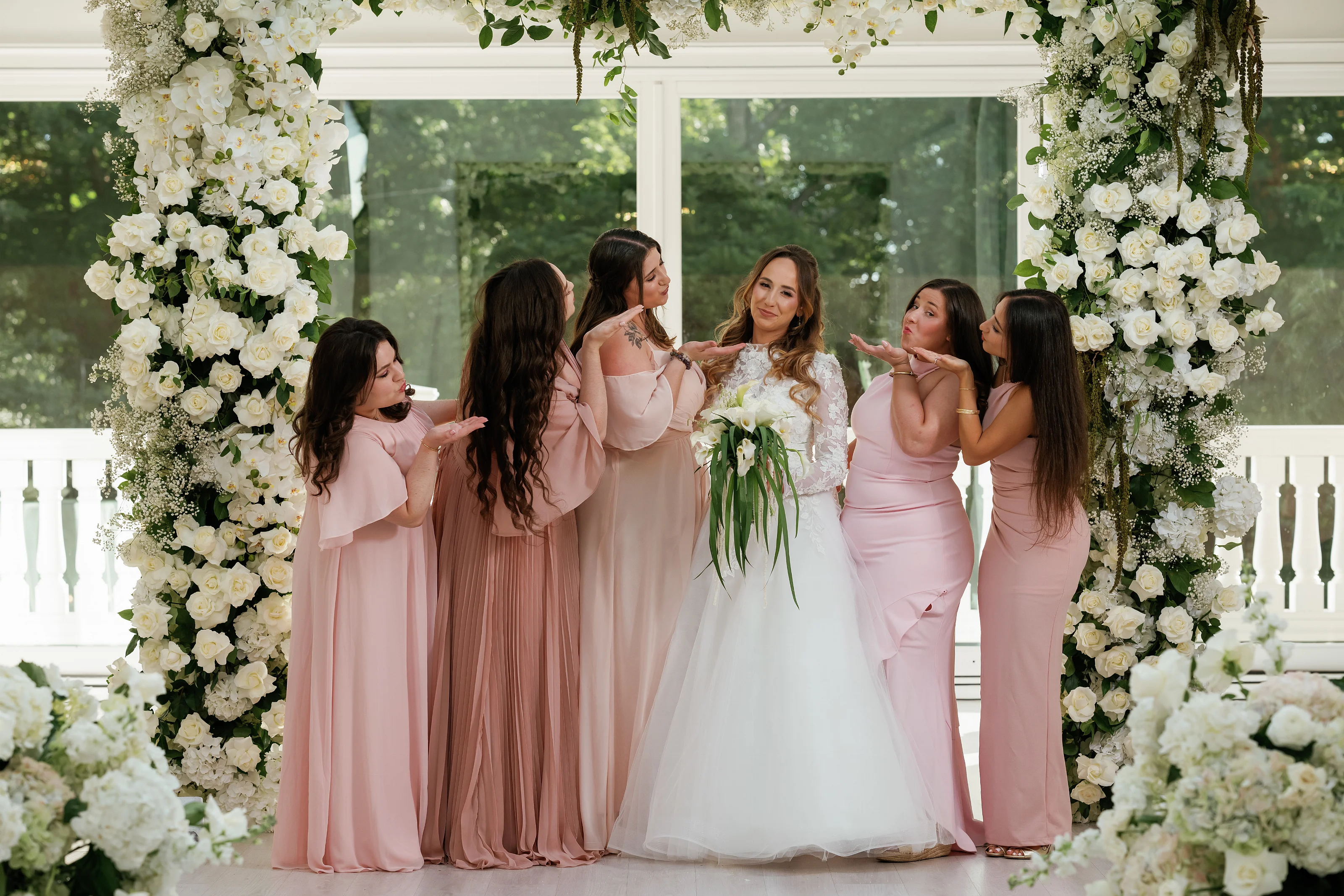Bride centered under a white floral canopy while bridesmaids in blush dresses surround her, gesturing and celebrating at a ceremony setting.