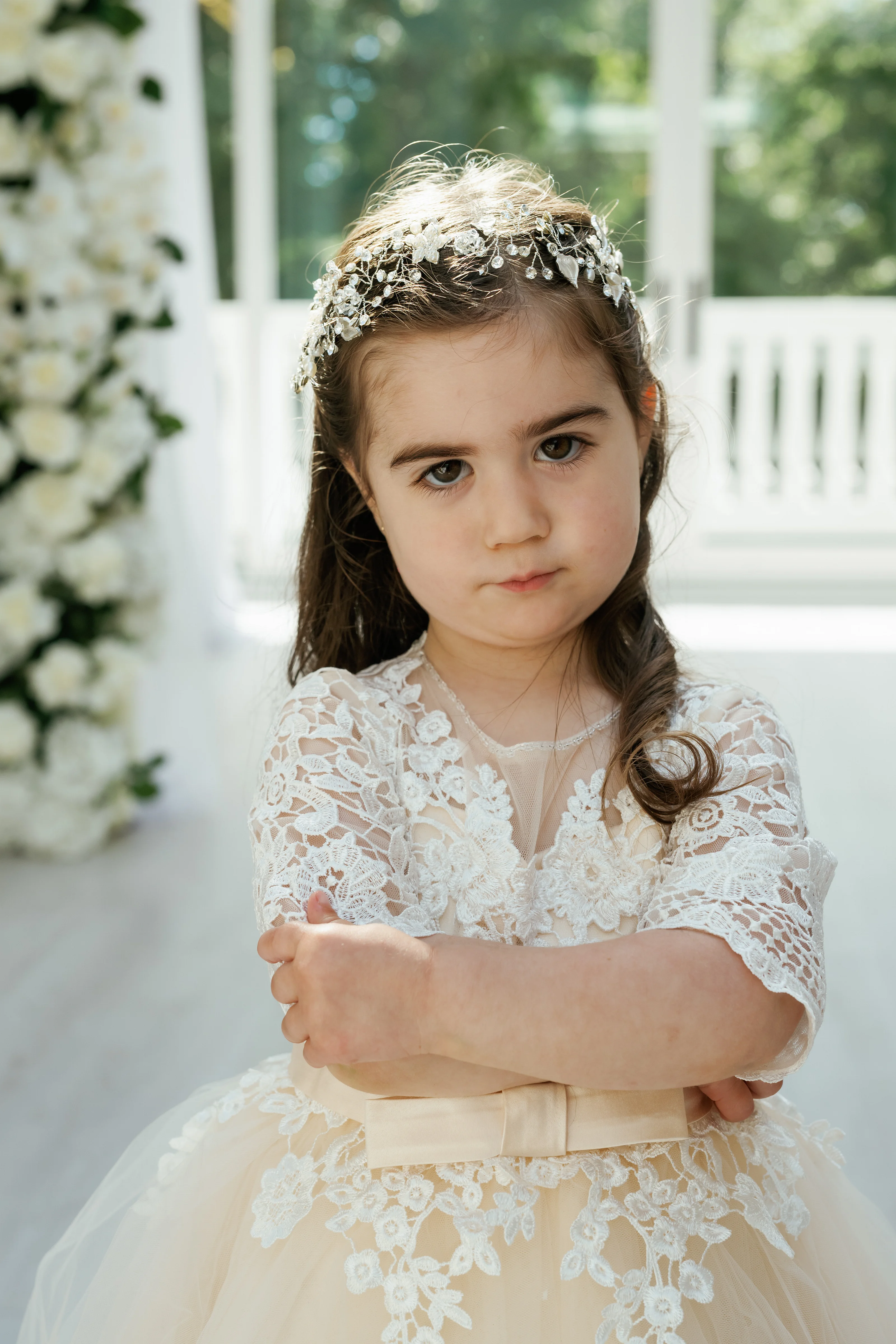 Portrait of a young girl in a white lace dress and floral headpiece standing indoors in soft daylight with arms crossed.