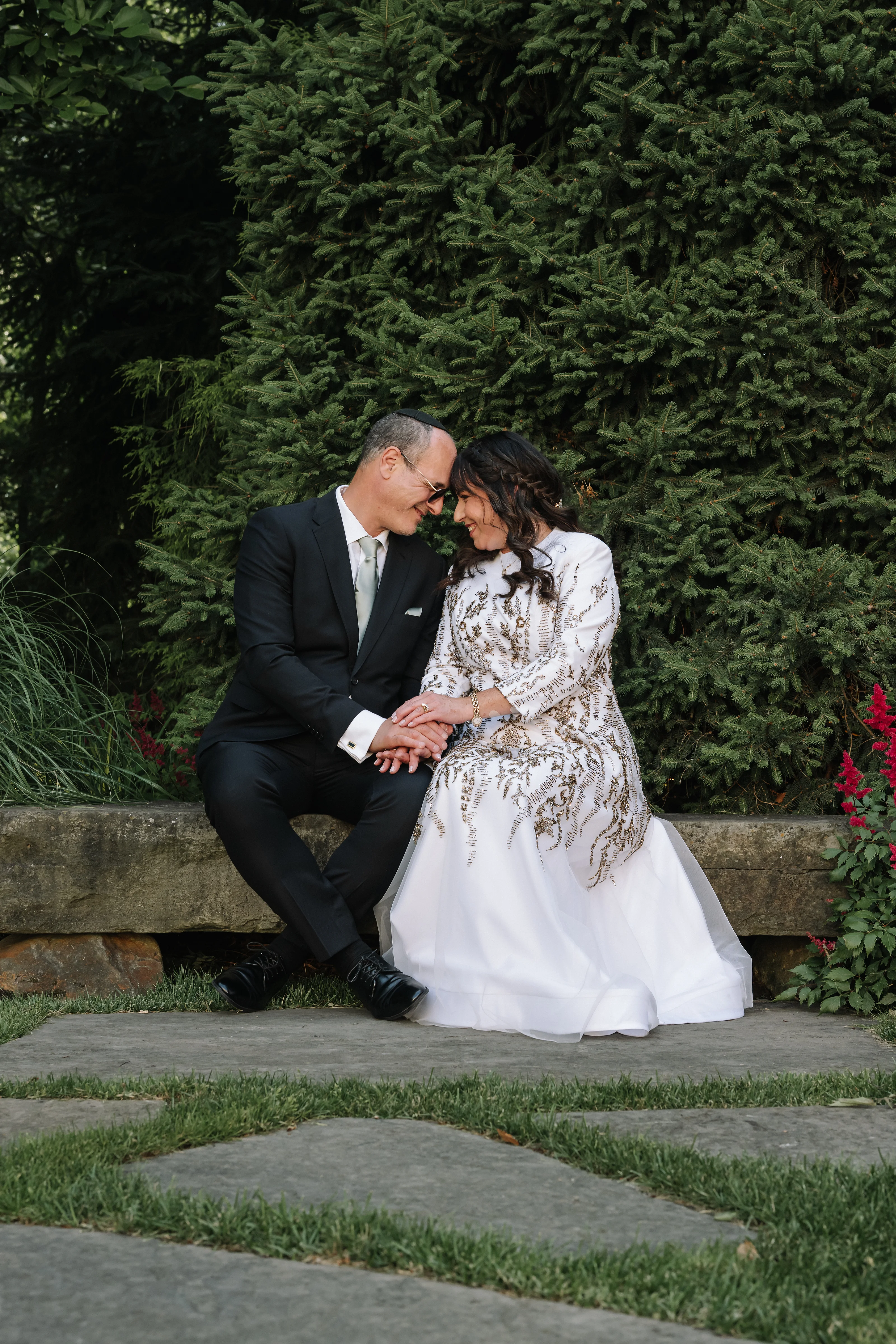 Bride and groom seated close together with foreheads touching, sharing an intimate moment outdoors against evergreen greenery.