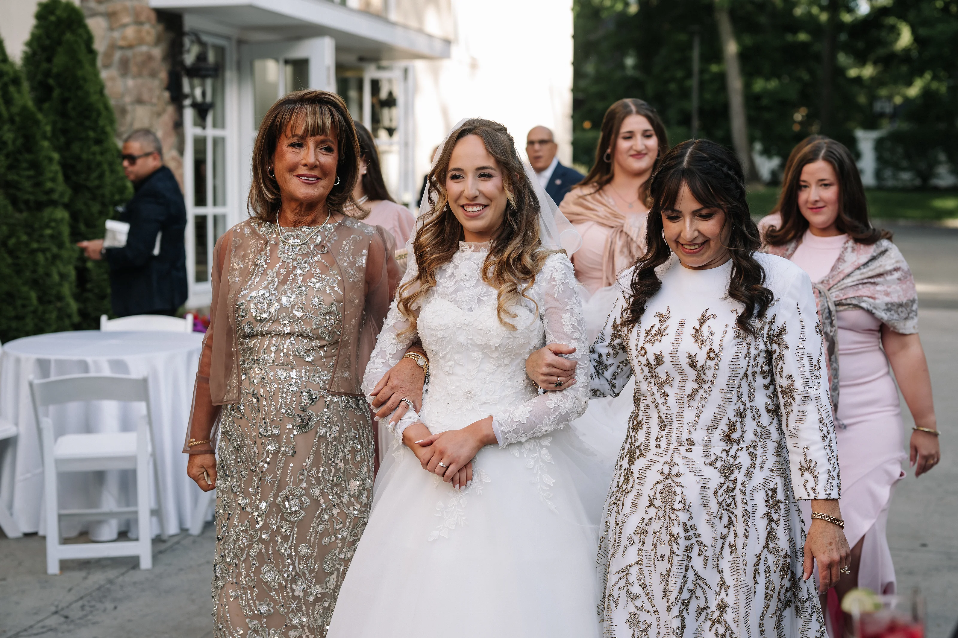 Bride in a white gown and veil smiling while walking with close family members at an outdoor wedding gathering, with guests in pastel attire behind her.