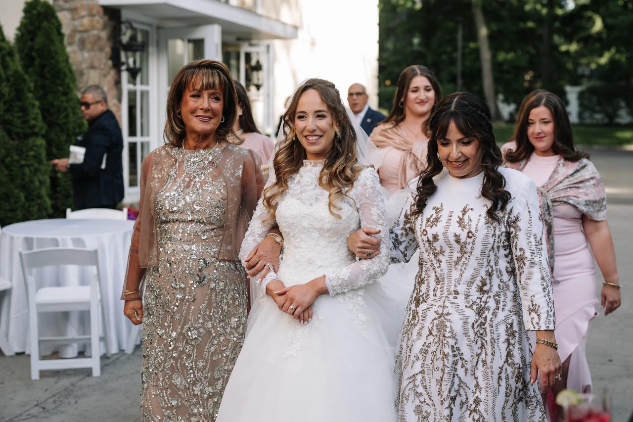 Bride in a white gown and veil smiling while walking with close family members at an outdoor wedding gathering, with guests in pastel attire behind her. Wedding at Crystal Plaza, Livingston, NJ.