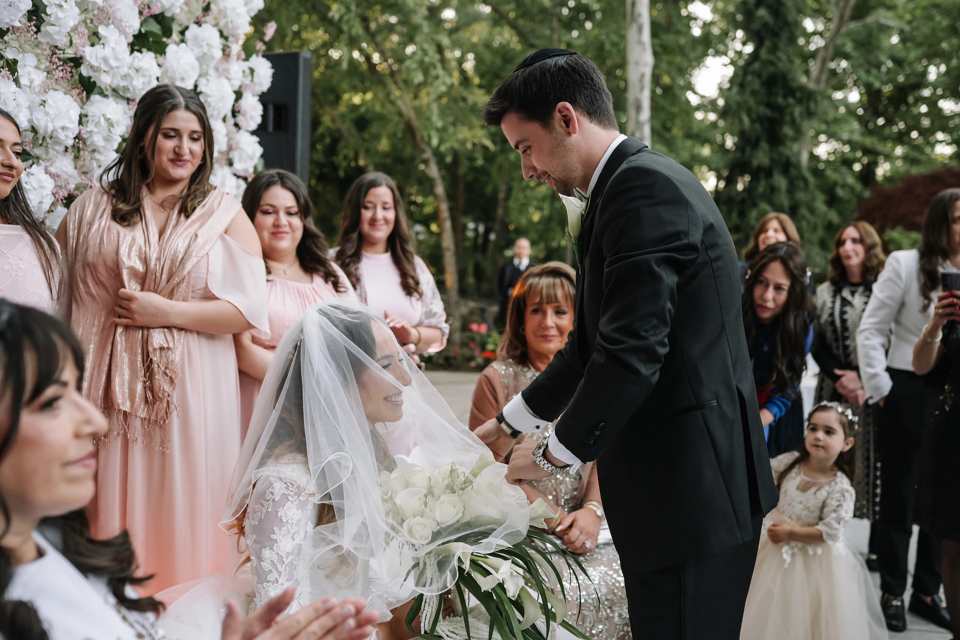 Groom leaning in during the badeken as the bride is covered, with family and a floral arch in the background.