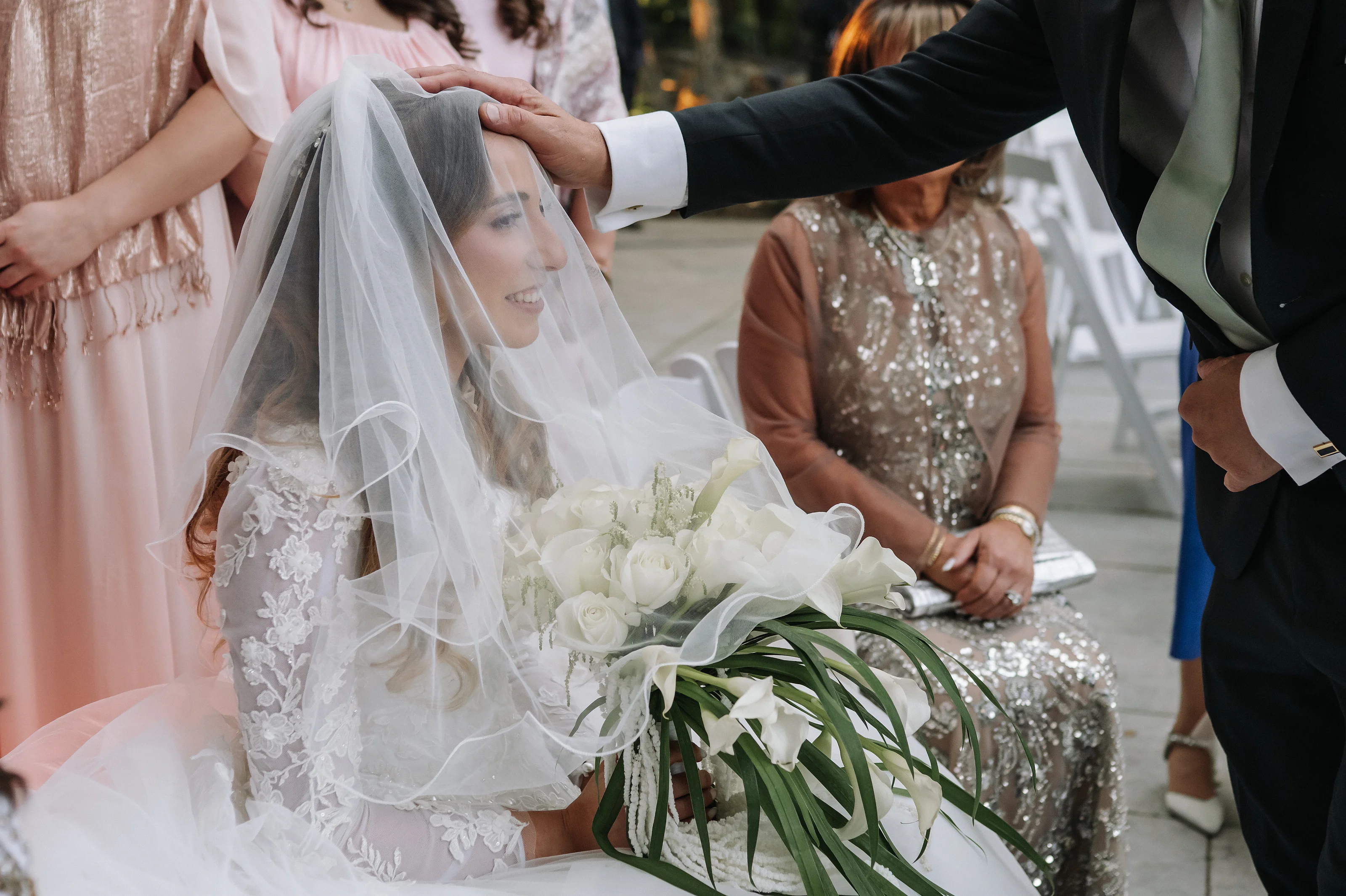 Bride smiling under a veil while the groom adjusts it during the badeken, with seated guests behind her.