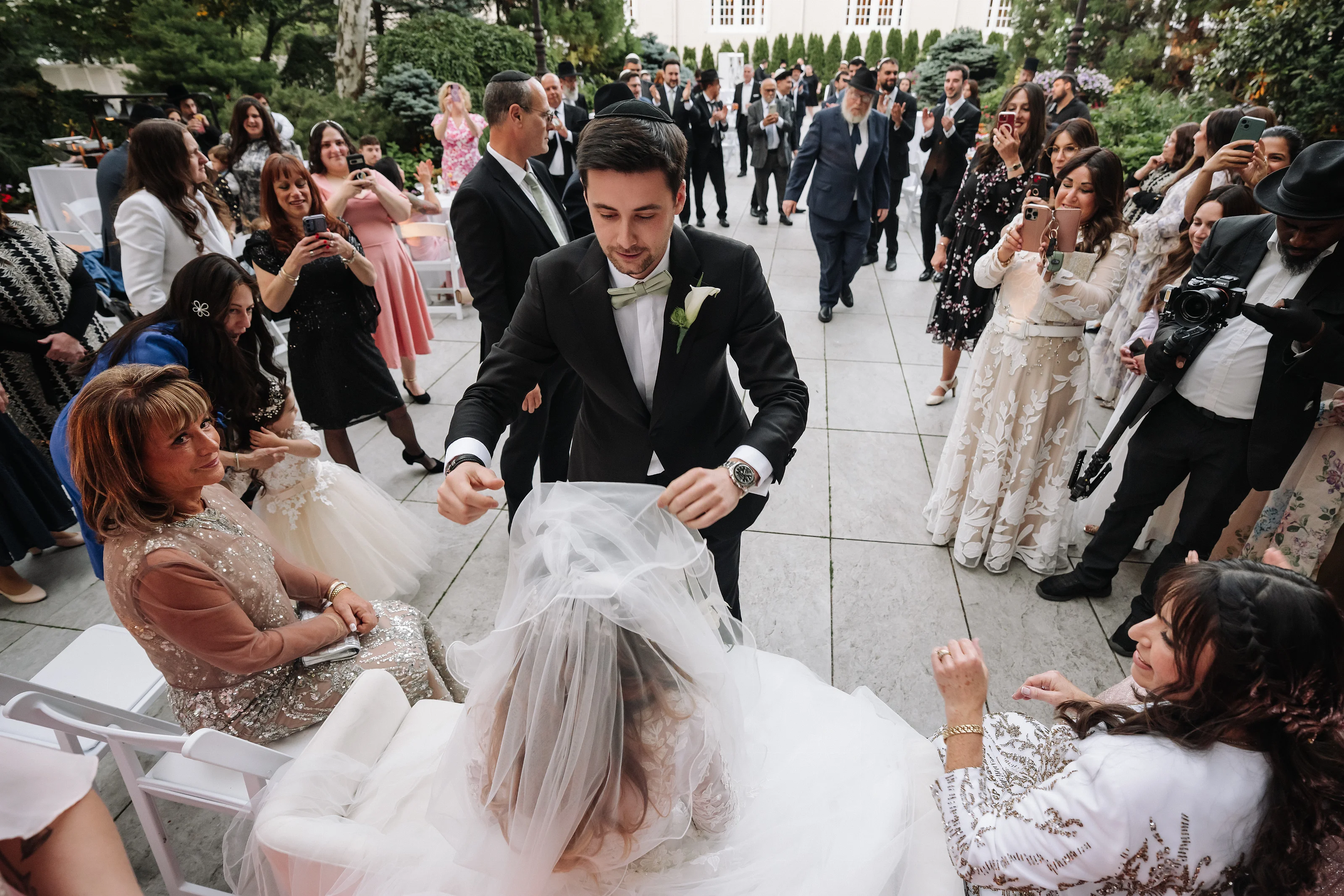 Groom bending forward smiling while guests clap during a wedding celebration outdoors along a garden walkway.